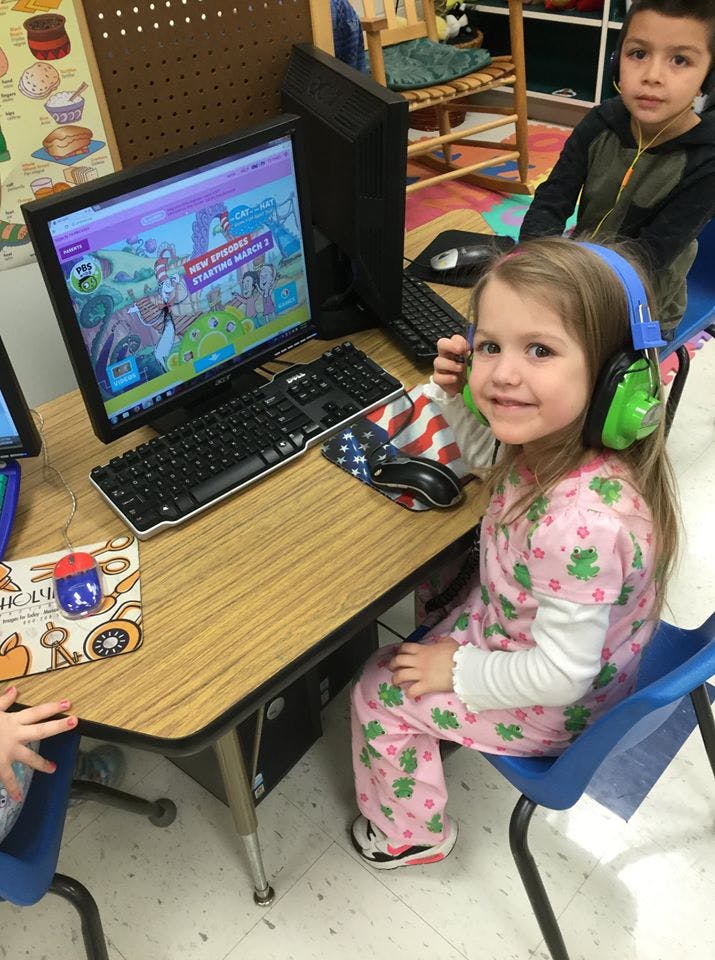 Child wearing headphones at a computer, another child in the background
