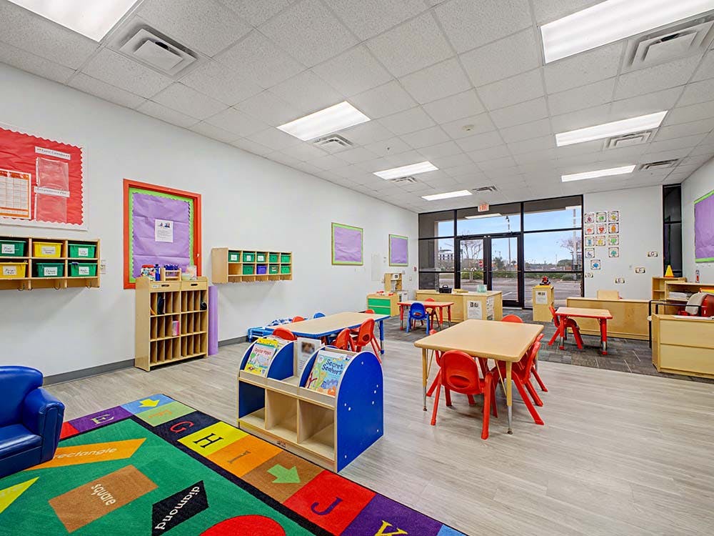 Empty classroom with small tables and chairs, shelves, and a colorful rug