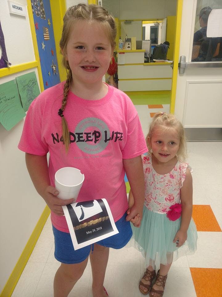 Two children standing, one holding a cup and paper, a paper with "May 25, 2018"
