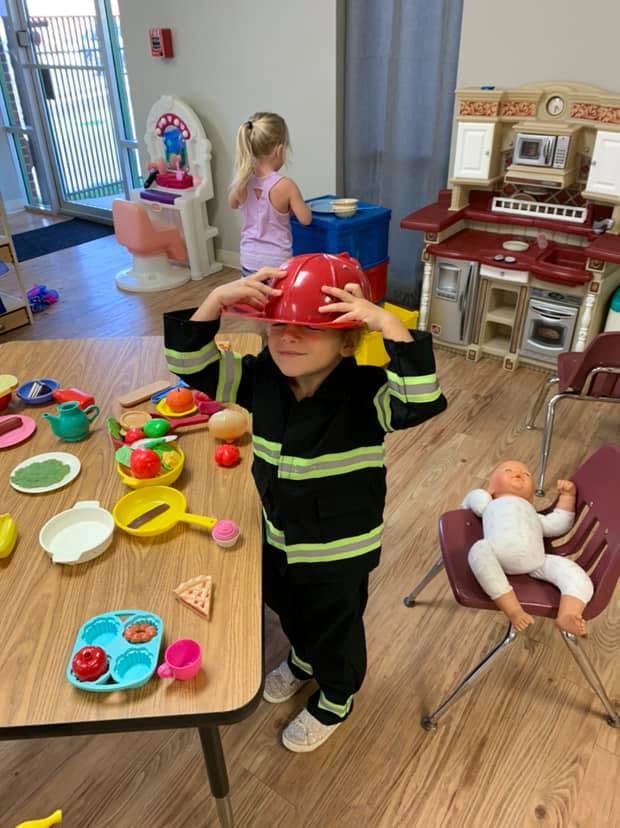 Child in a firefighter costume wearing a red helmet, standing by a table with toys