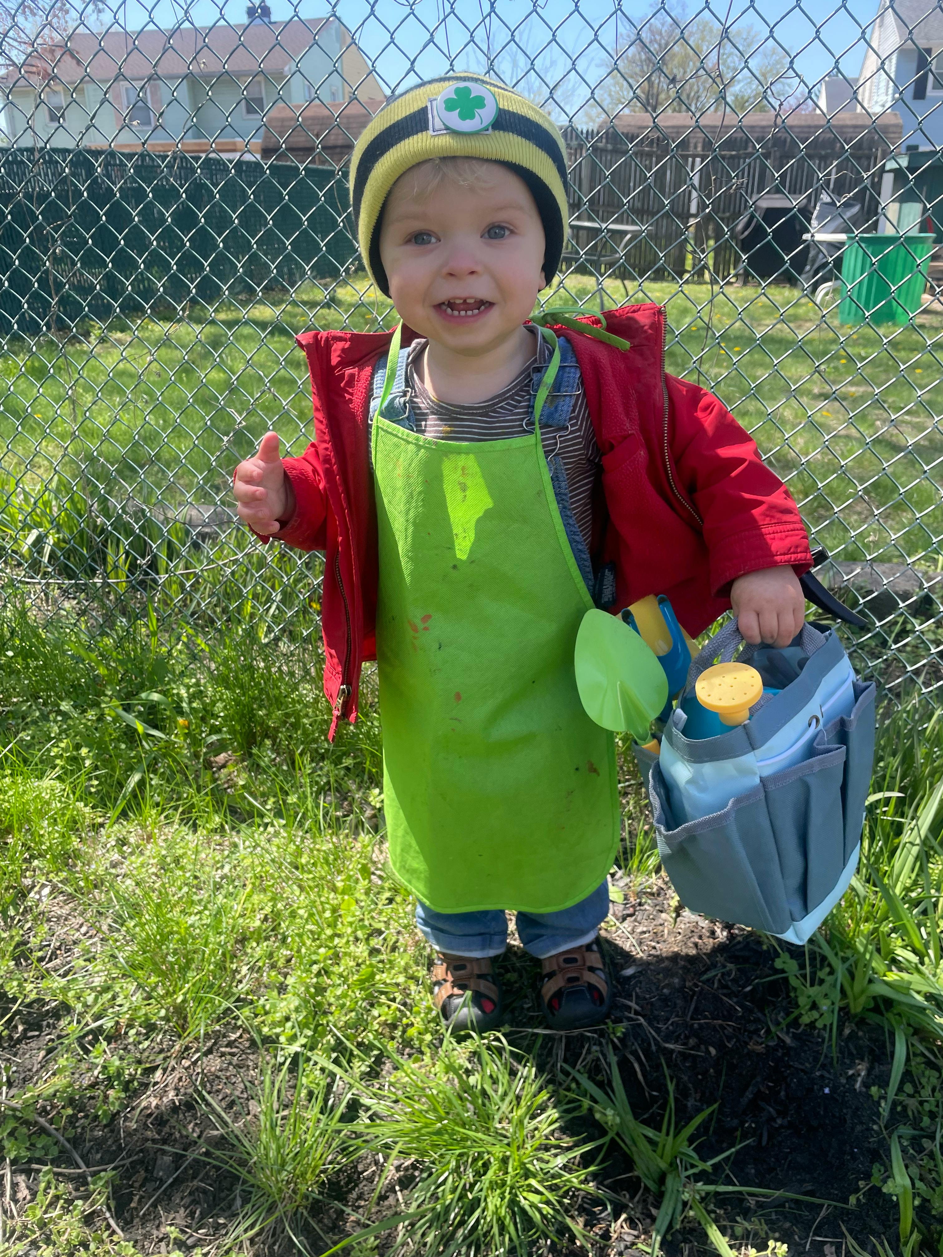 Child in a green apron and red jacket holding a gardening tool bag