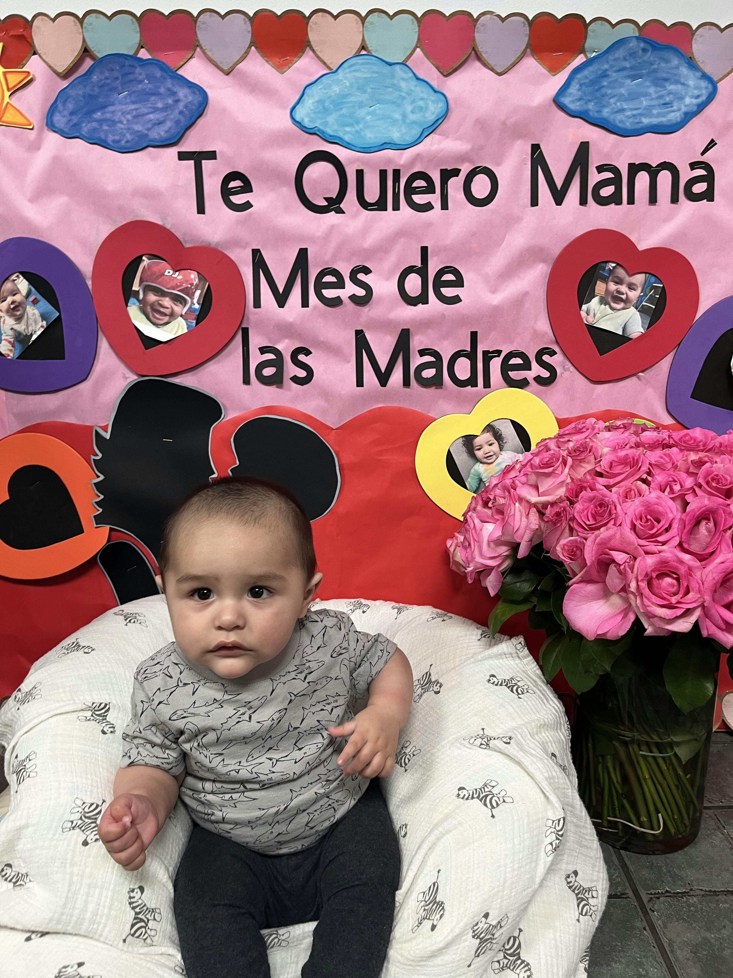 Baby sitting in front of a pink banner with text and a vase of pink roses