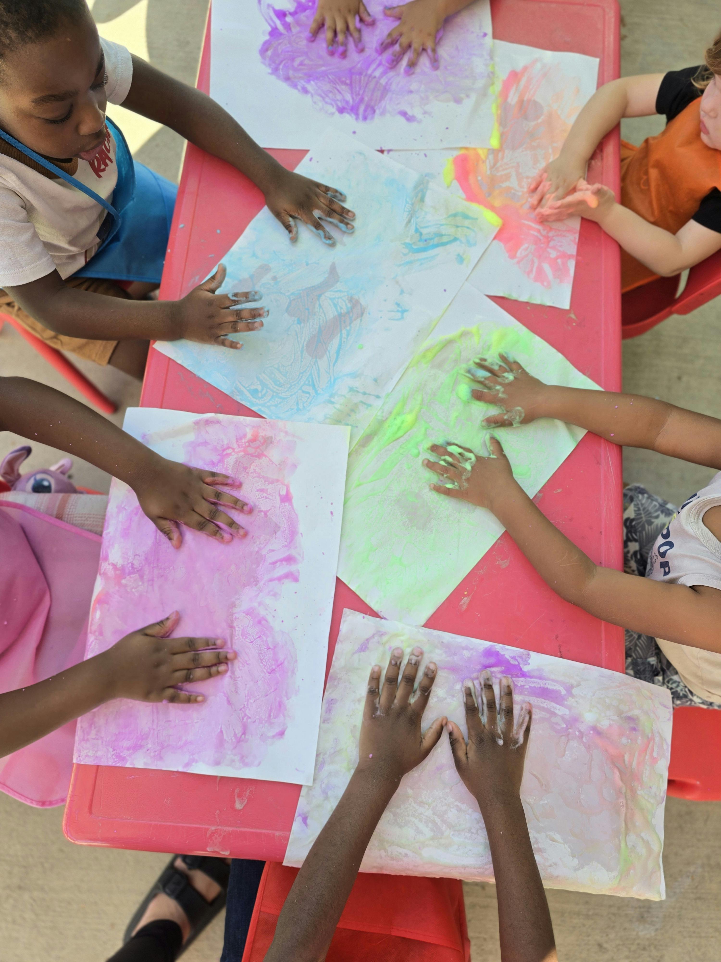 Children using their hands to paint on sheets of paper at a red table