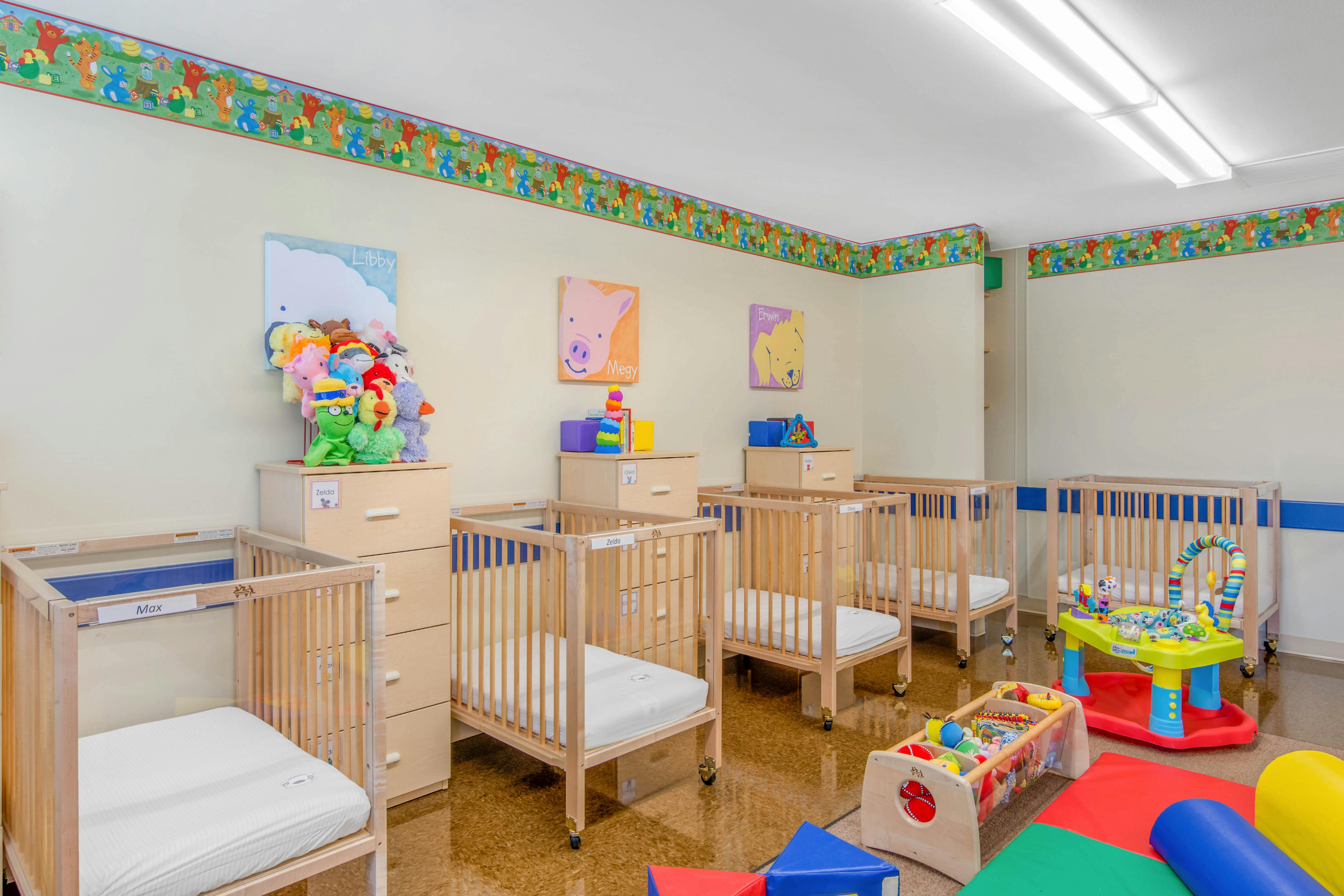 A room with several cribs, dressers with stuffed animals, and infant play equipment