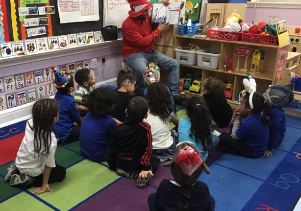 Person in red hat reading a book to children sitting on a colorful rug