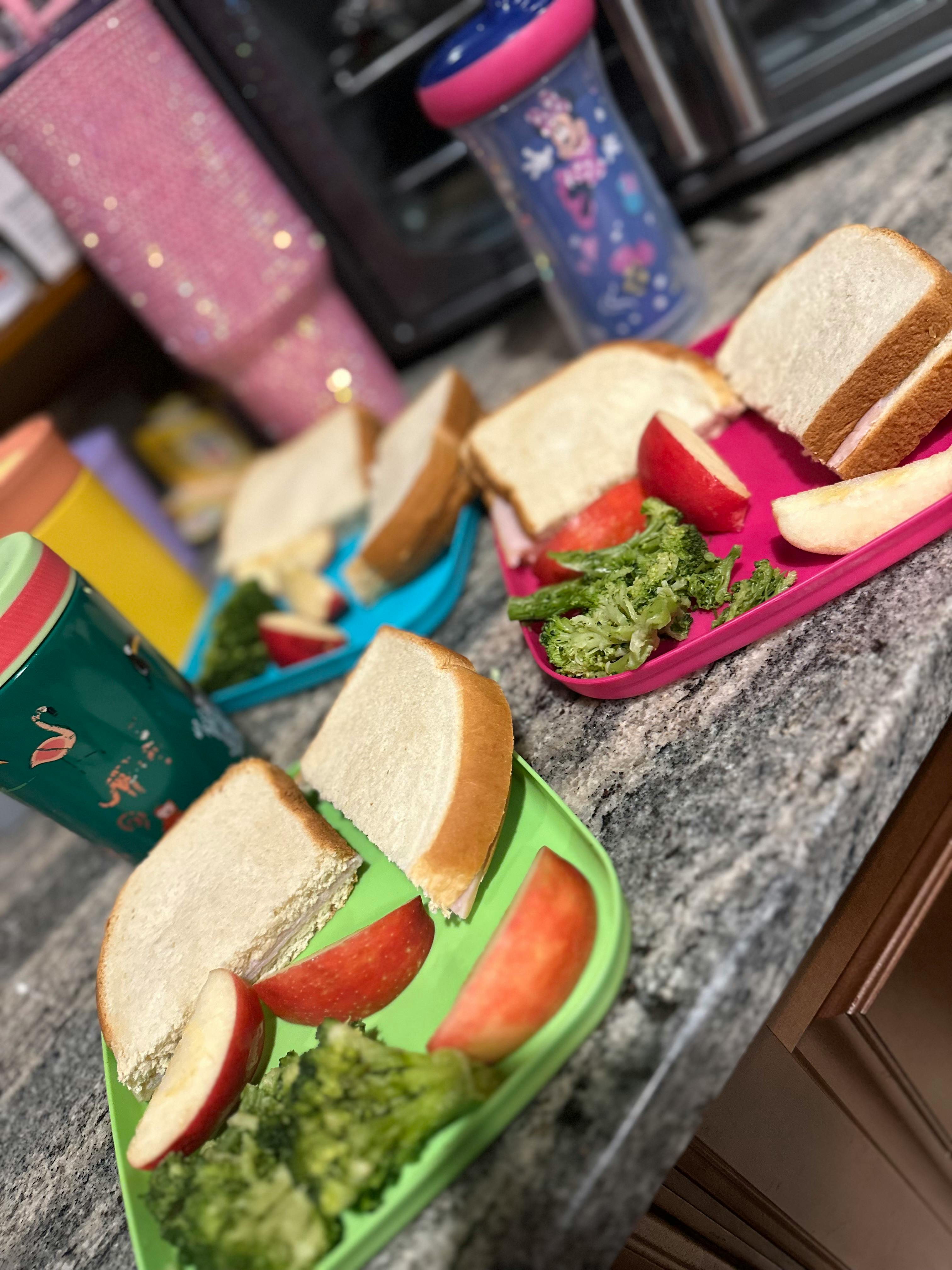 Multiple plates with sandwiches, apple slices, and broccoli