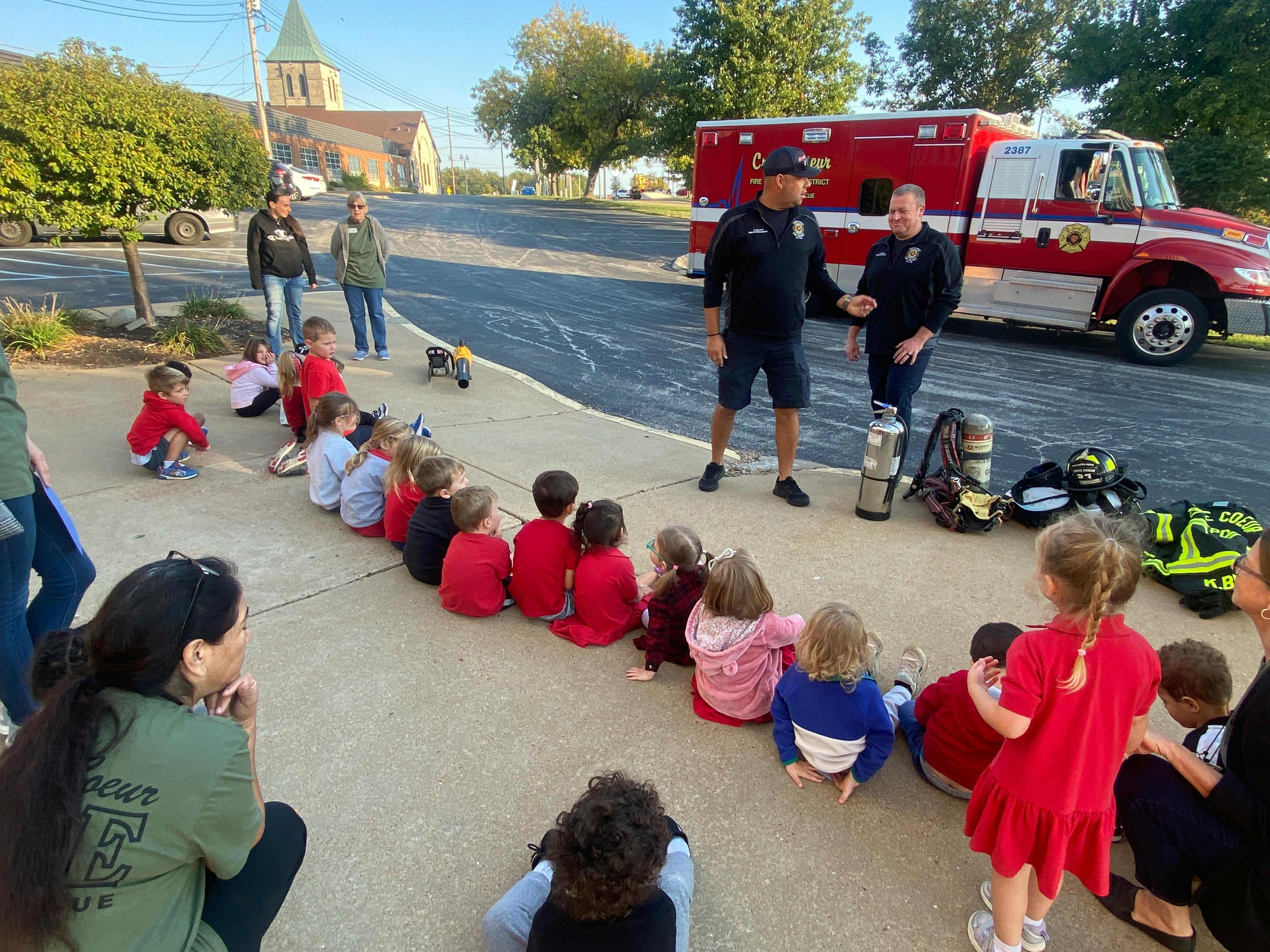 Children sitting on ground listening to two people in uniform near a fire truck
