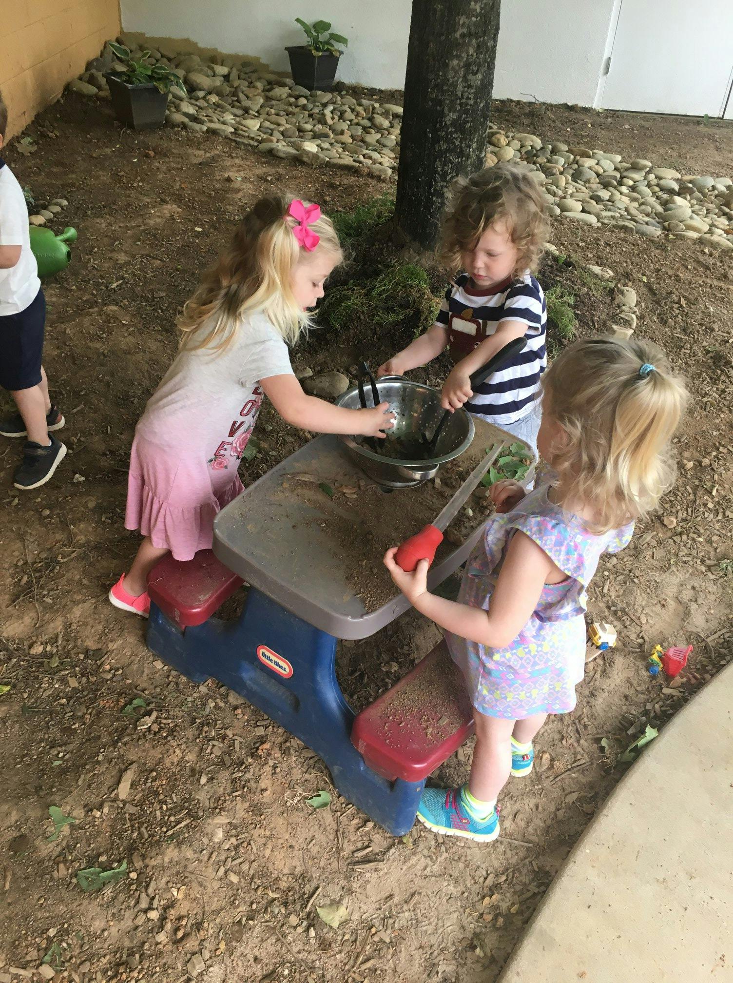 Three children playing with a bowl and utensils at a small picnic table outdoors