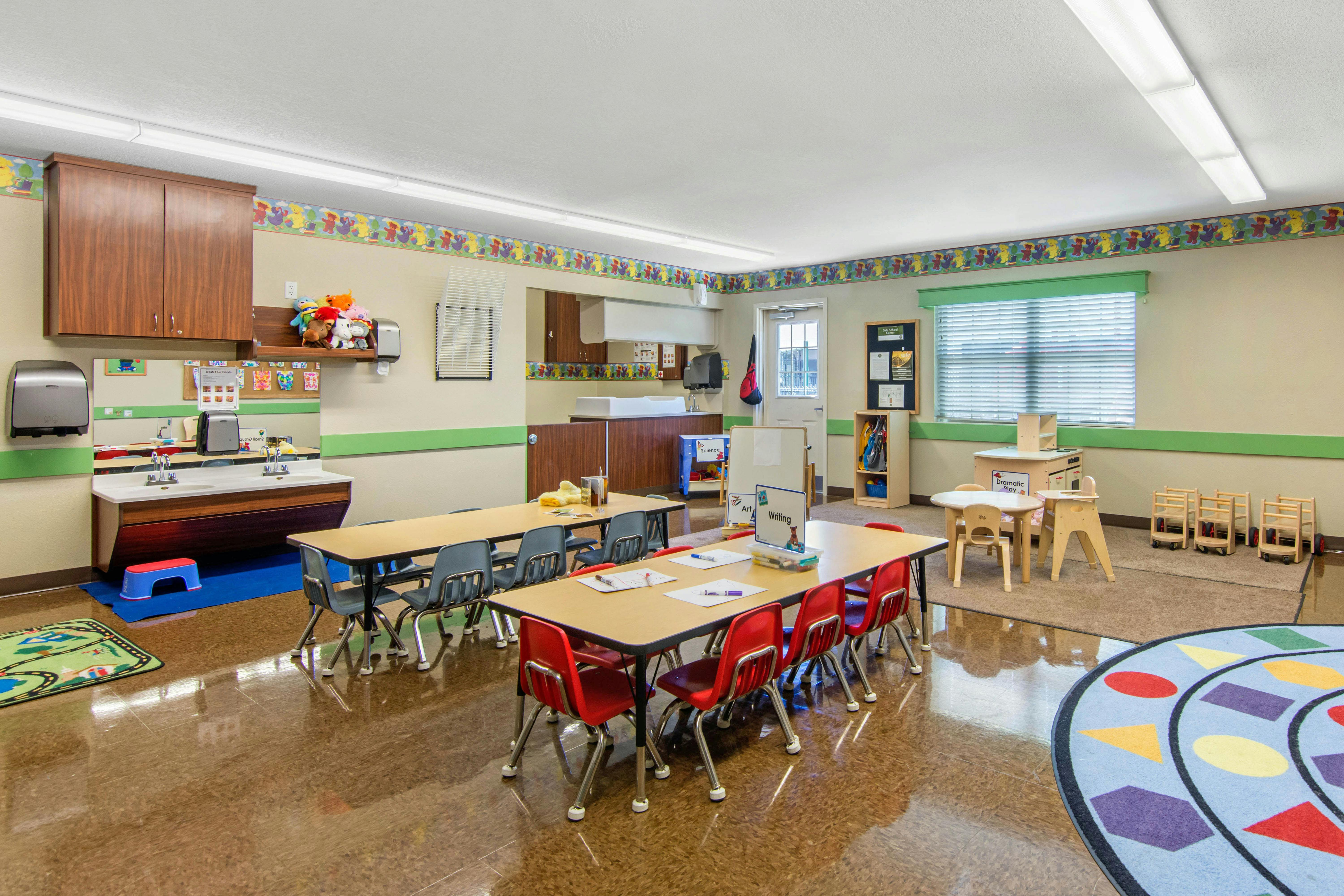 An empty classroom with child-sized tables and chairs, a sink area, and various play stations