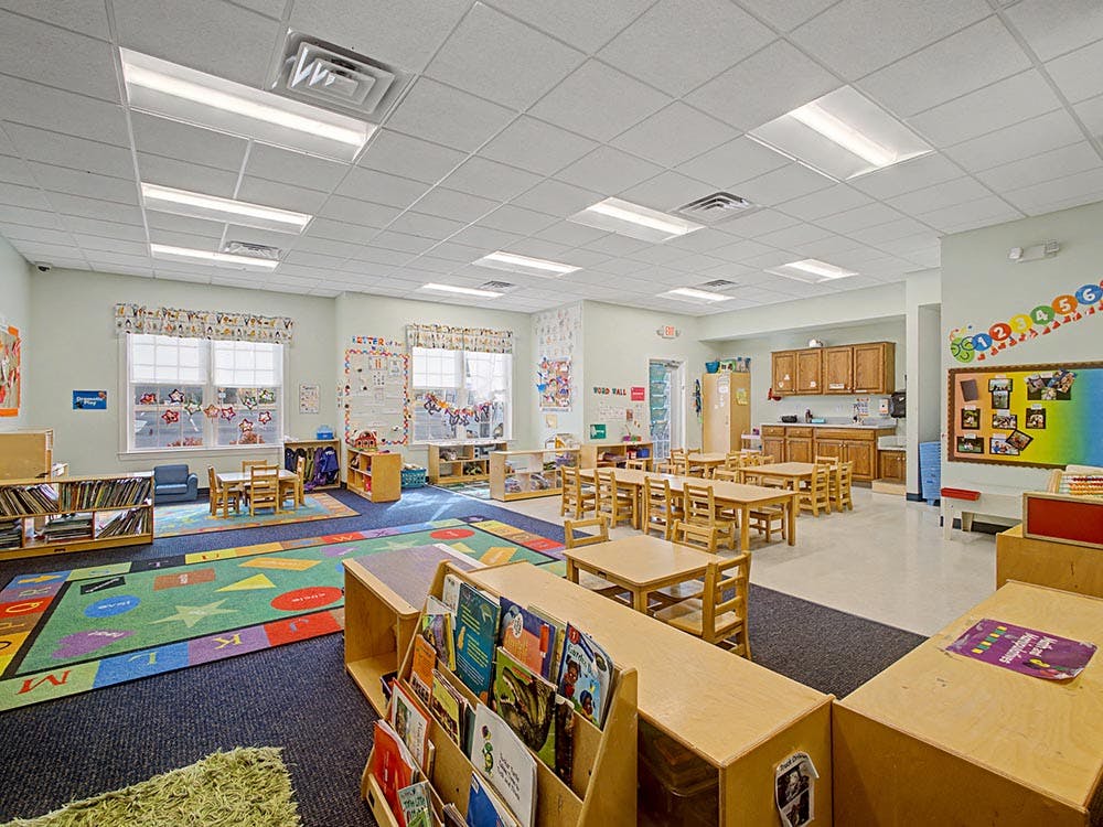 A colorful classroom with small tables and chairs, shelves, and a rug with letters and shapes