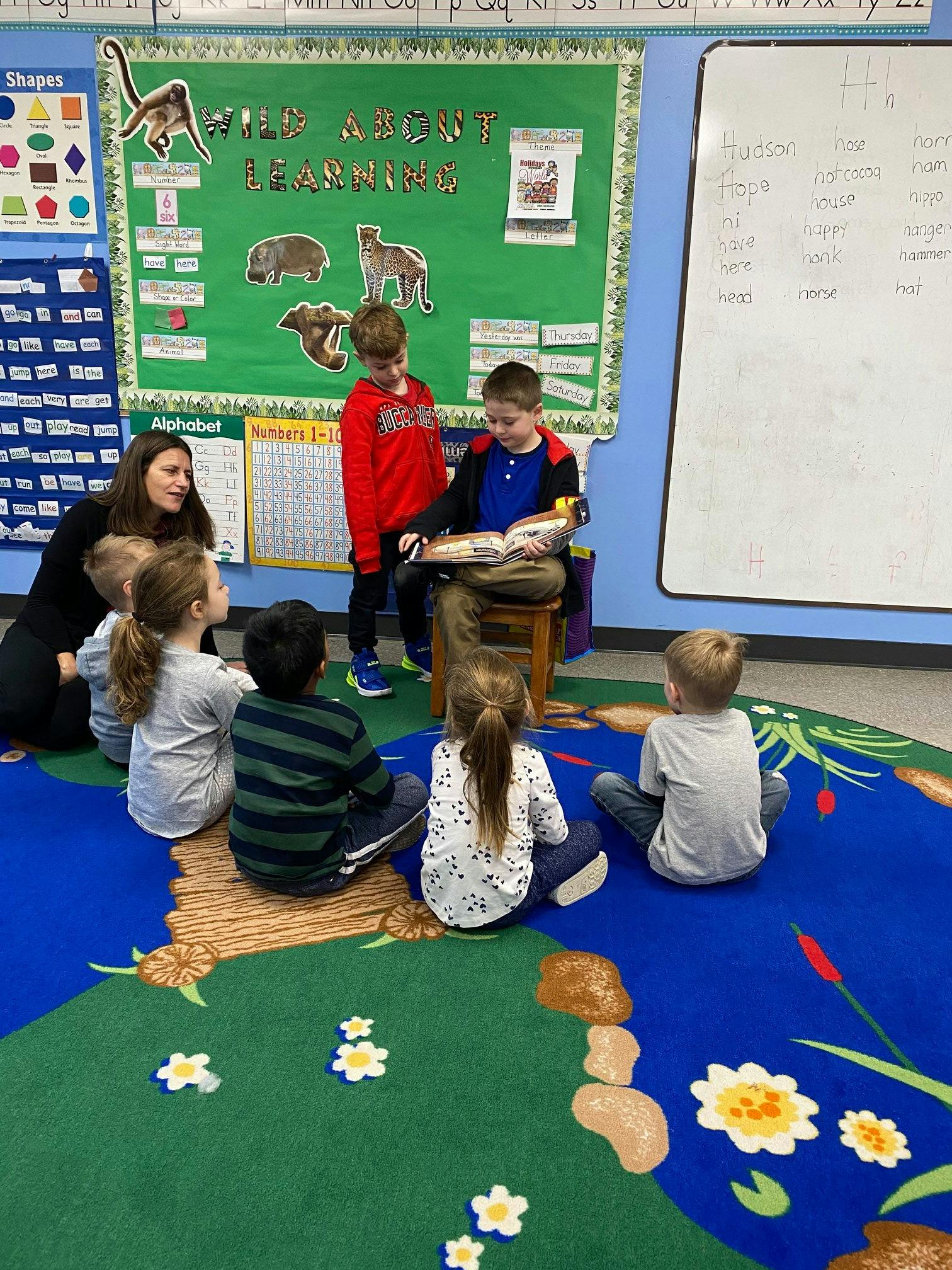 Adult and children sitting on a rug, one child reading a book aloud
