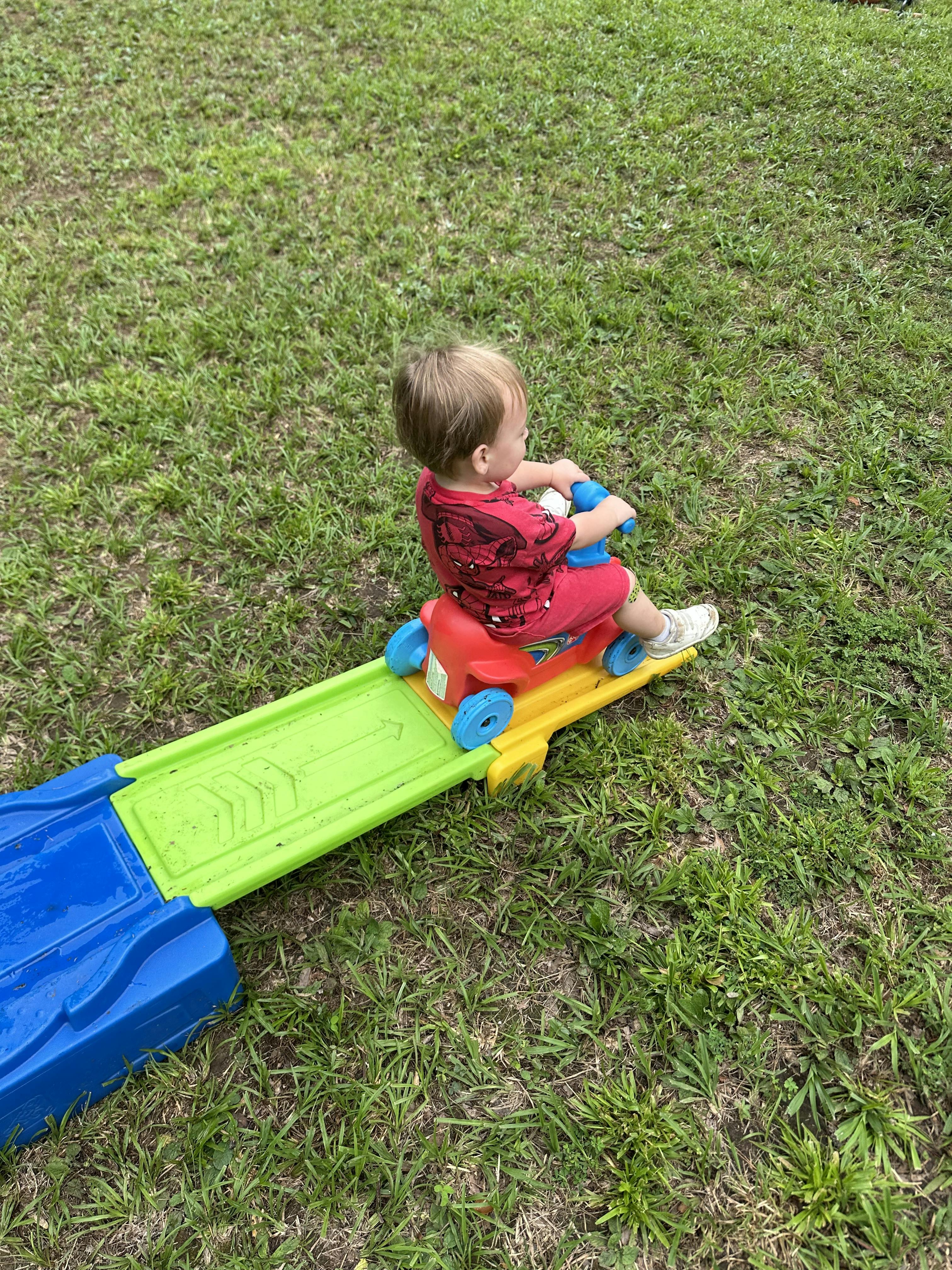 Child riding a toy car on a ramp in a grassy area