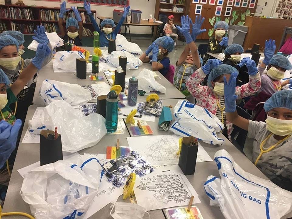 Children in masks, hairnets, and gloves raising hands at a table with supplies