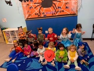 Children in costumes sitting on a mat in a room with a spider decoration