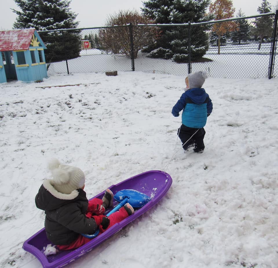 Child sitting in a purple sled while another child walks in the snow