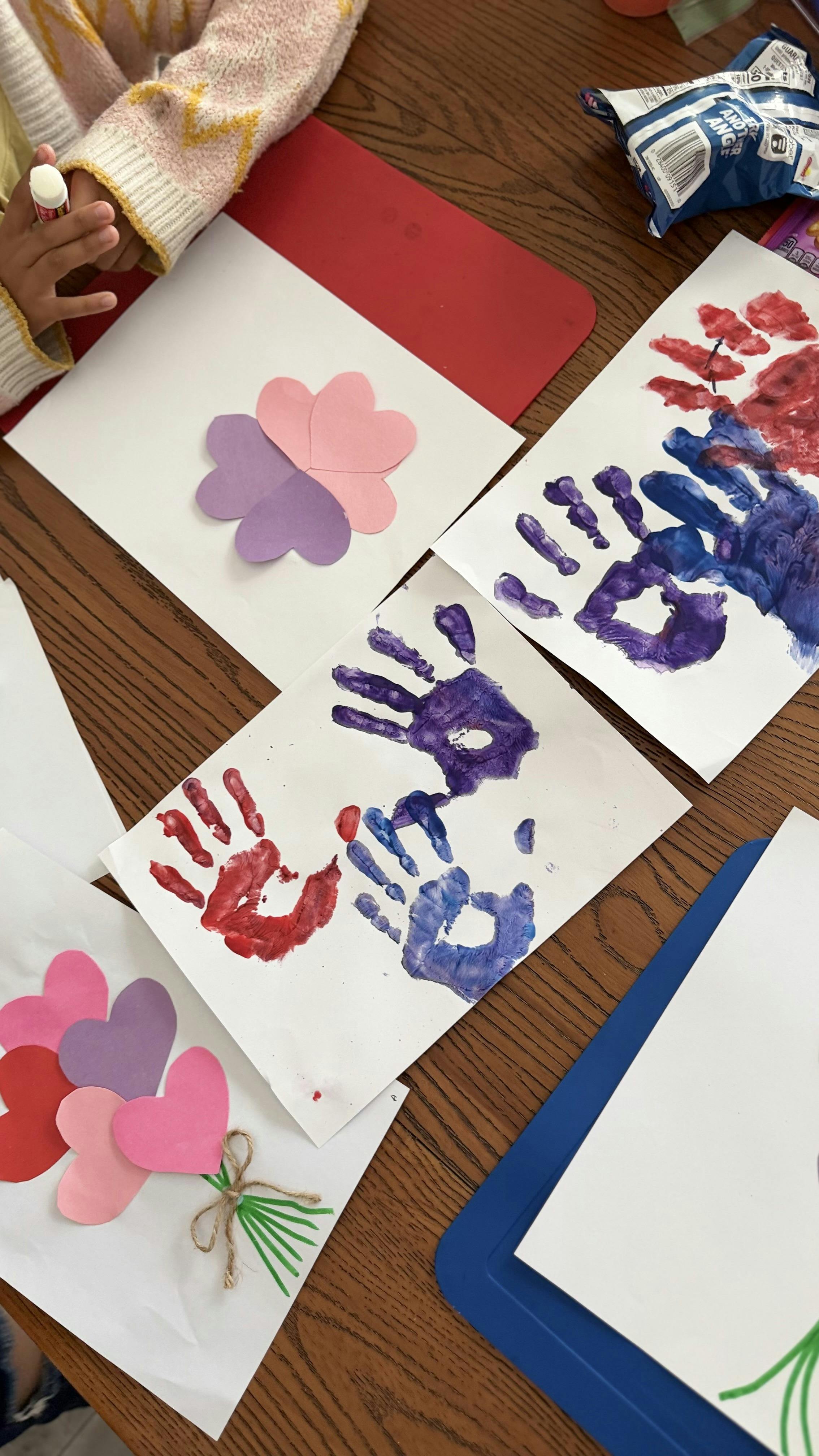 Child crafting with paper hearts and glue stick, surrounded by handprint art