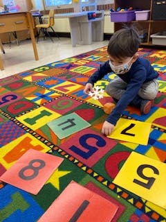 Child in mask playing with number squares on a colorful rug