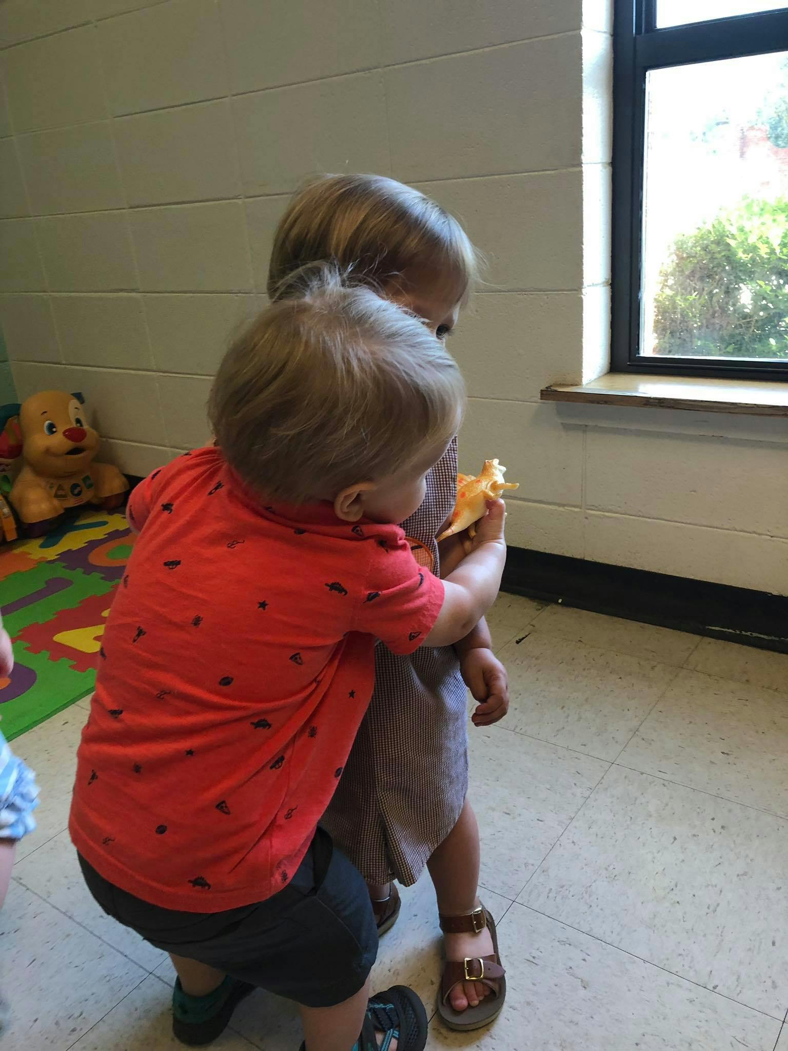 Child in red shirt hugging another child holding a toy