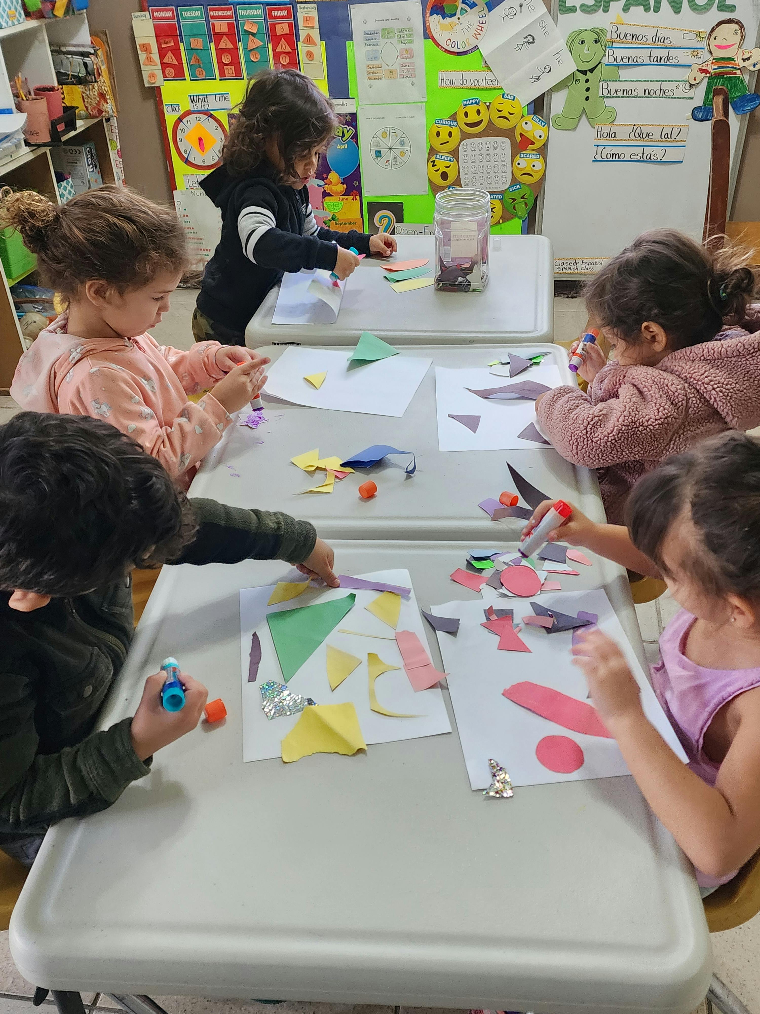 Children doing crafts at a table, gluing paper shapes onto white sheets