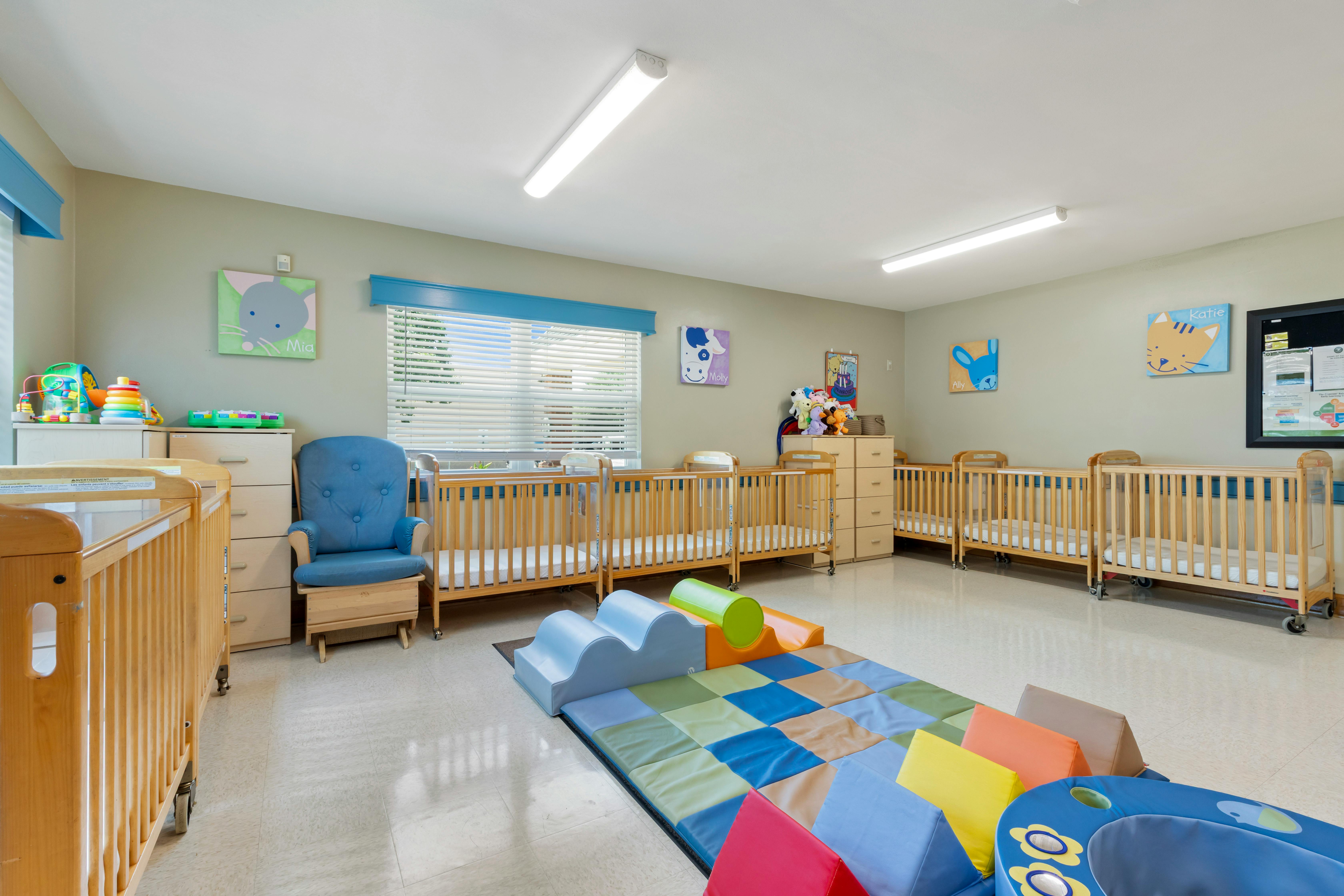 A brightly lit nursery room with several wooden cribs, a blue rocking chair, and colorful play mats on the floor