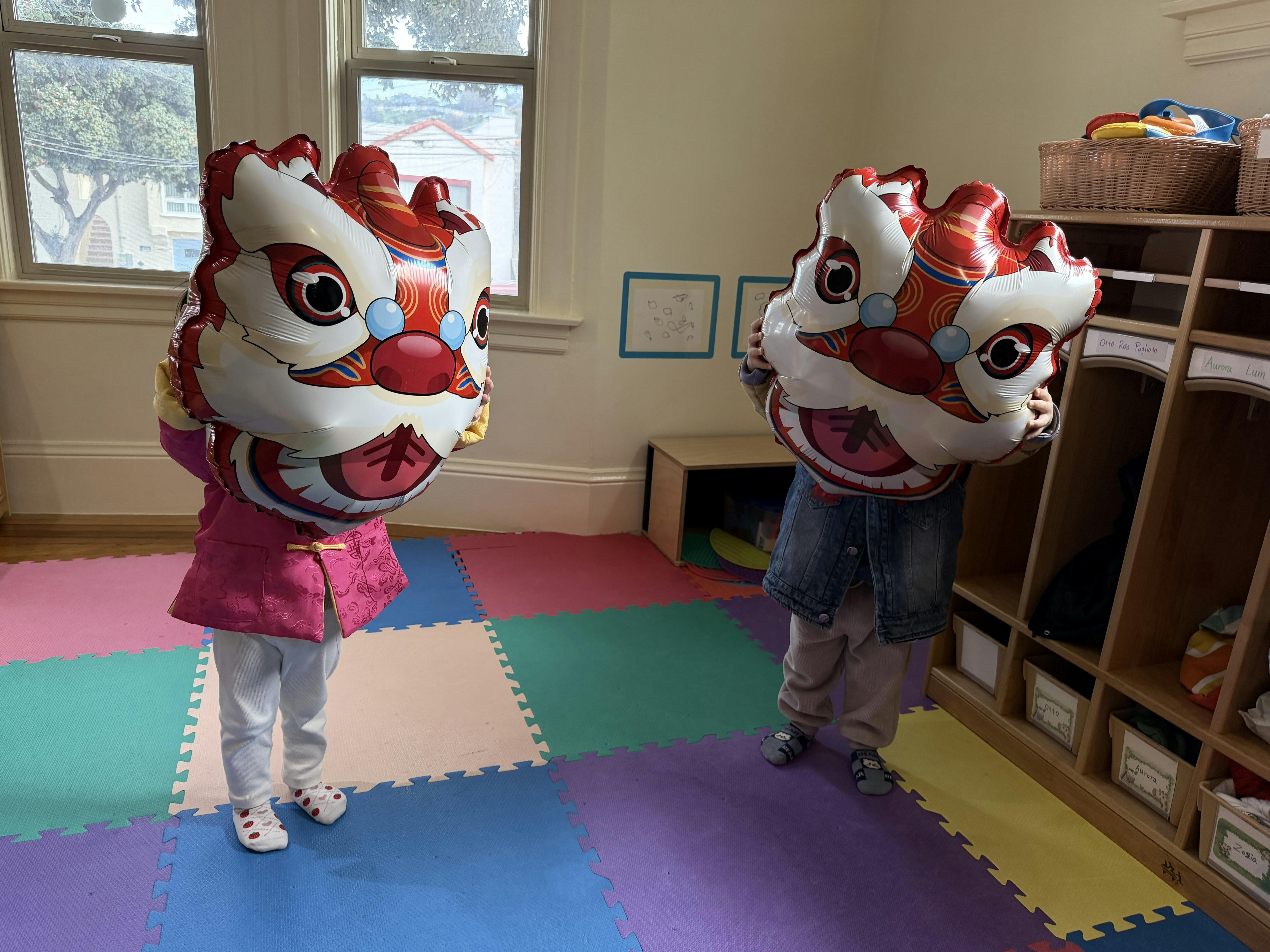 Two children holding up lion dance head balloons in a classroom