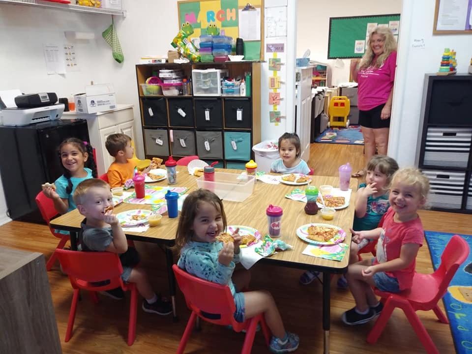 Children eating at a table in a classroom with an adult standing in the background