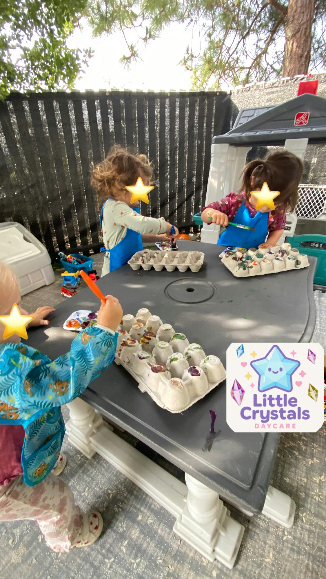 Three children painting egg cartons at a table outdoors