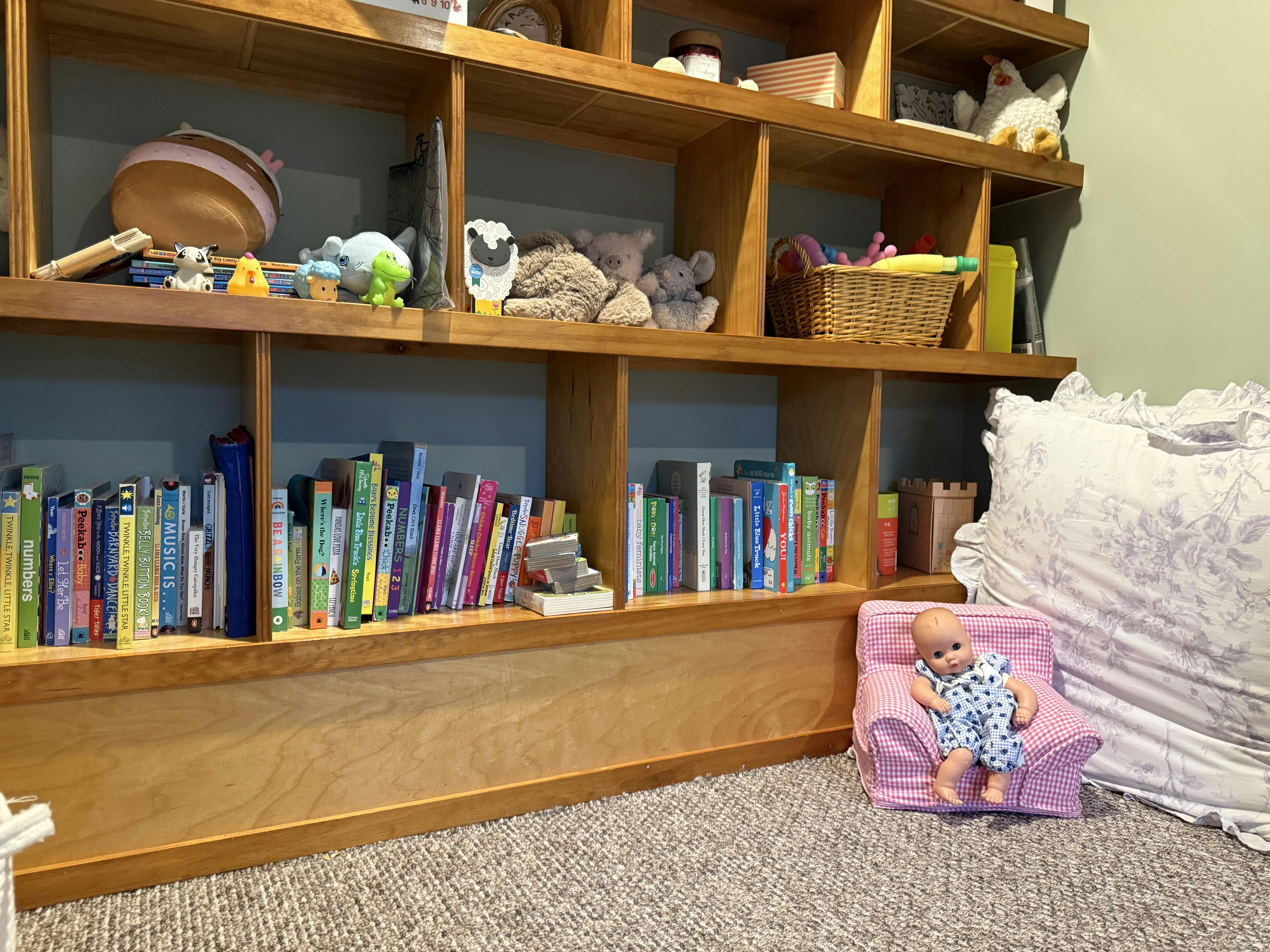 Doll in a doll chair next to a wooden bookshelf with books and toys