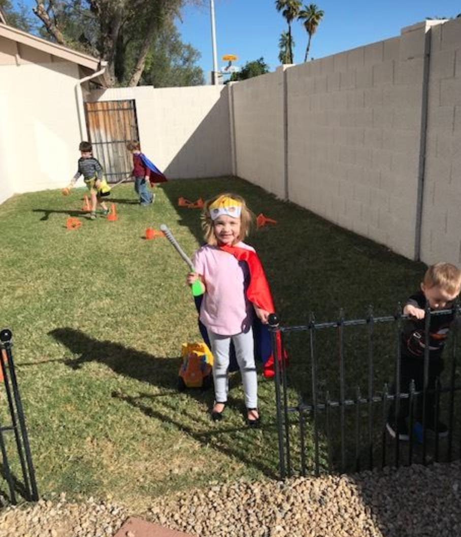Child in a cape holding a stick, with other children in a grassy yard