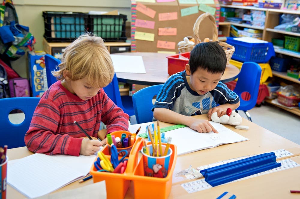 Two children drawing and writing at a table in a classroom