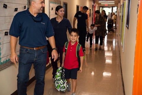 People walking down a school hallway, child in red shirt with backpack in foreground