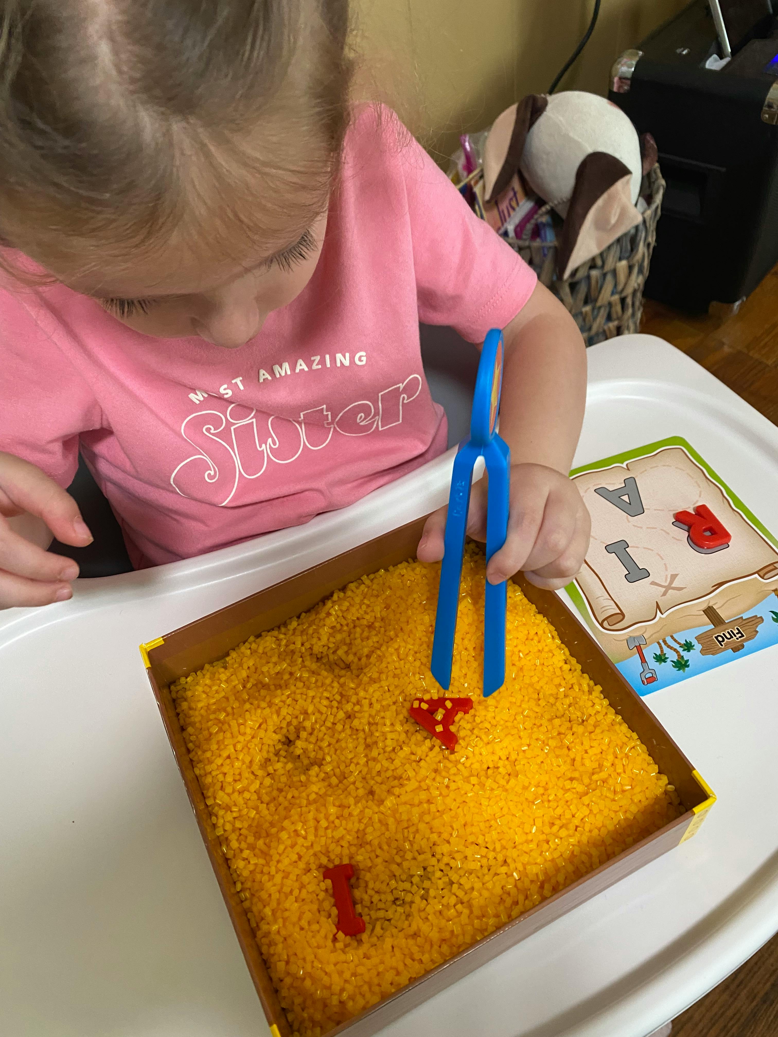 Child using blue tongs to pick up a red letter from a box of yellow beads