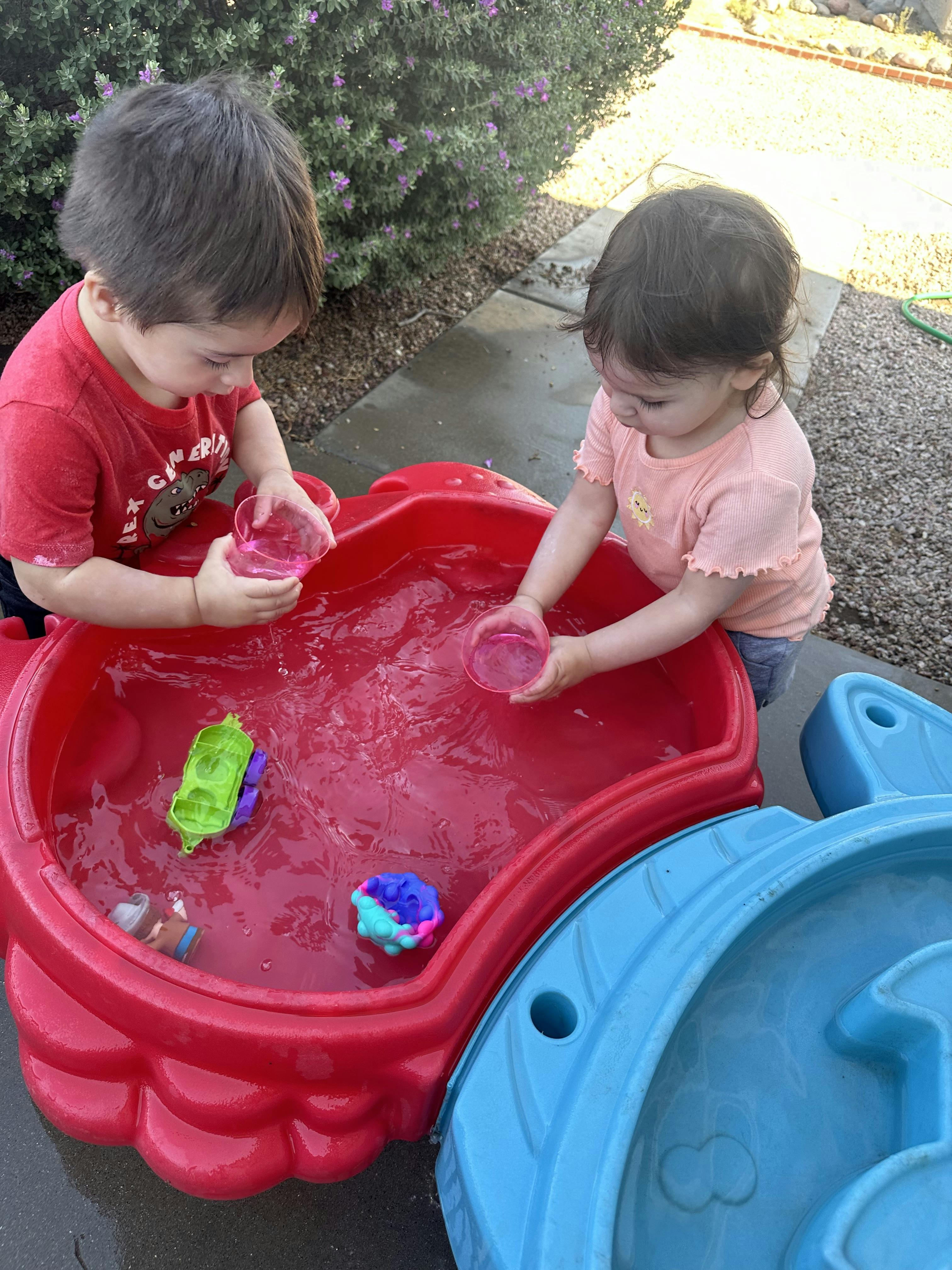 Two children playing with water and plastic cups in a red toy pool