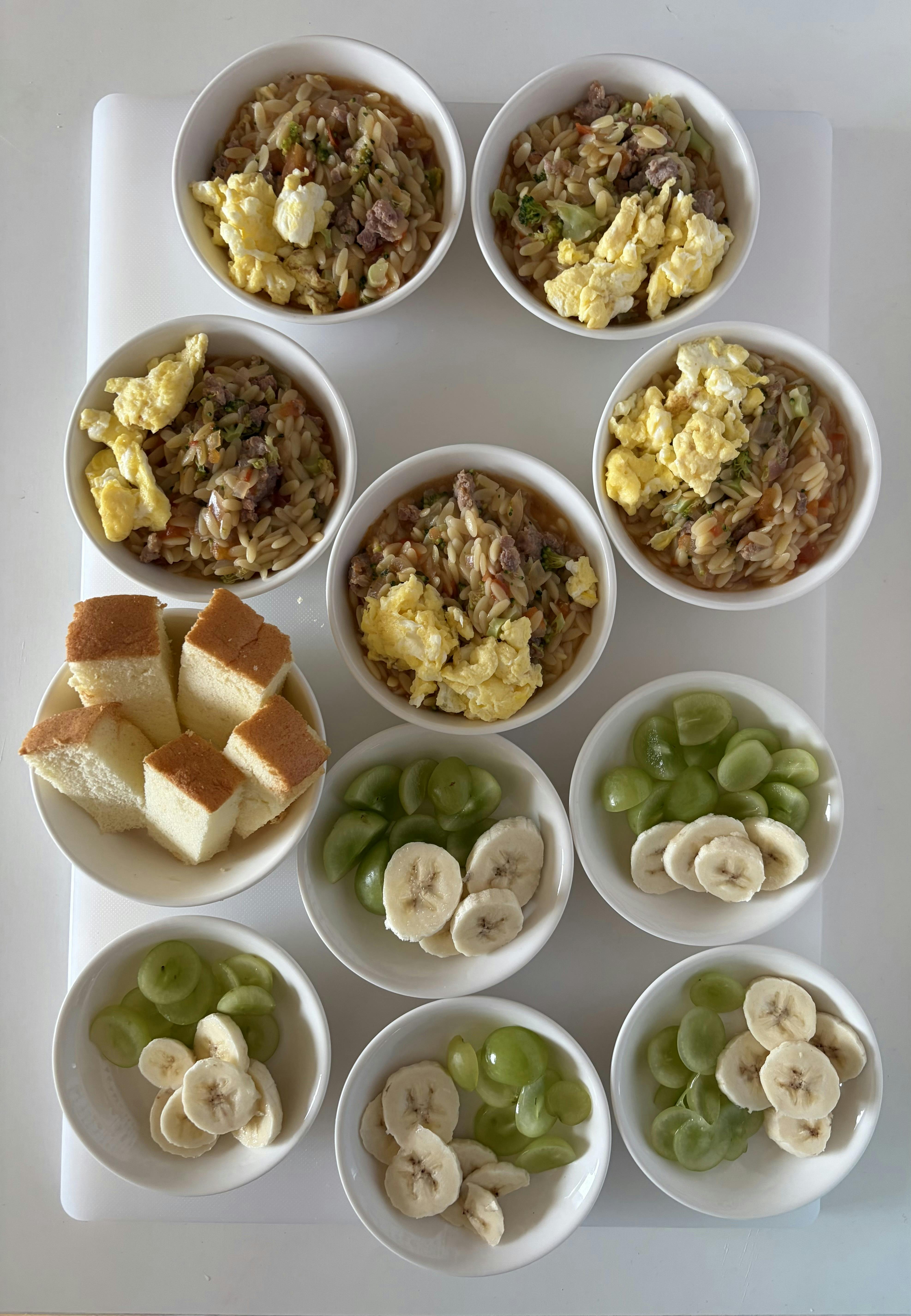 Several bowls of orzo with egg and smaller bowls of sliced fruit and bread