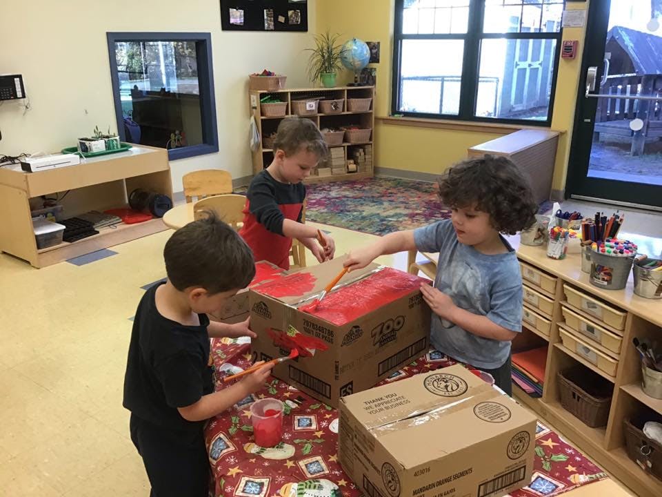 Three children painting cardboard boxes in a classroom