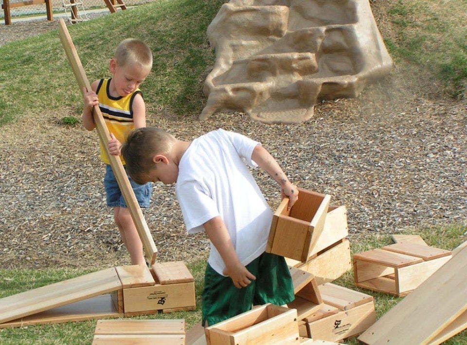 Two children playing with wooden blocks and a long stick outdoors
