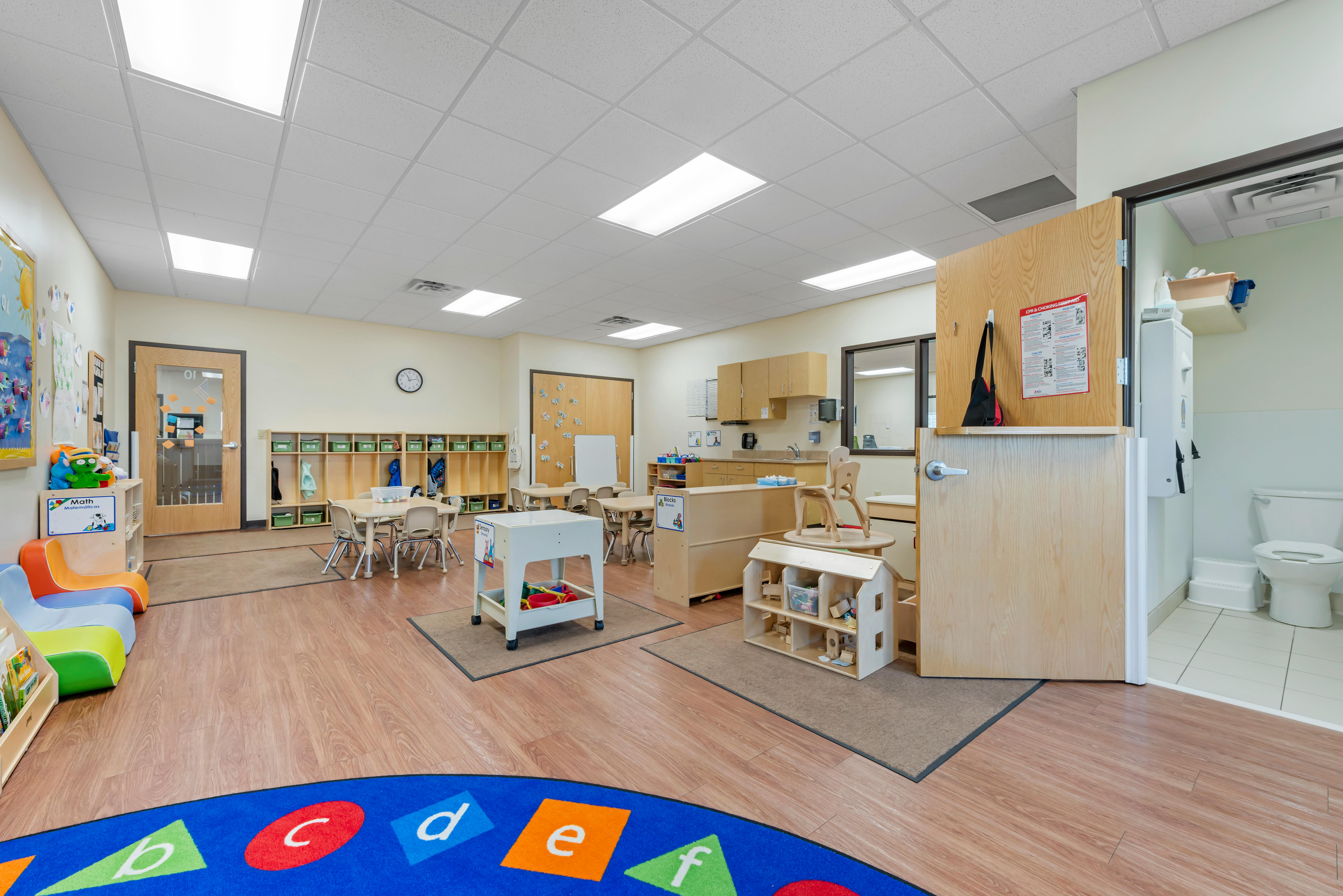 Classroom with child-sized tables and chairs, a play kitchen, and an open bathroom door