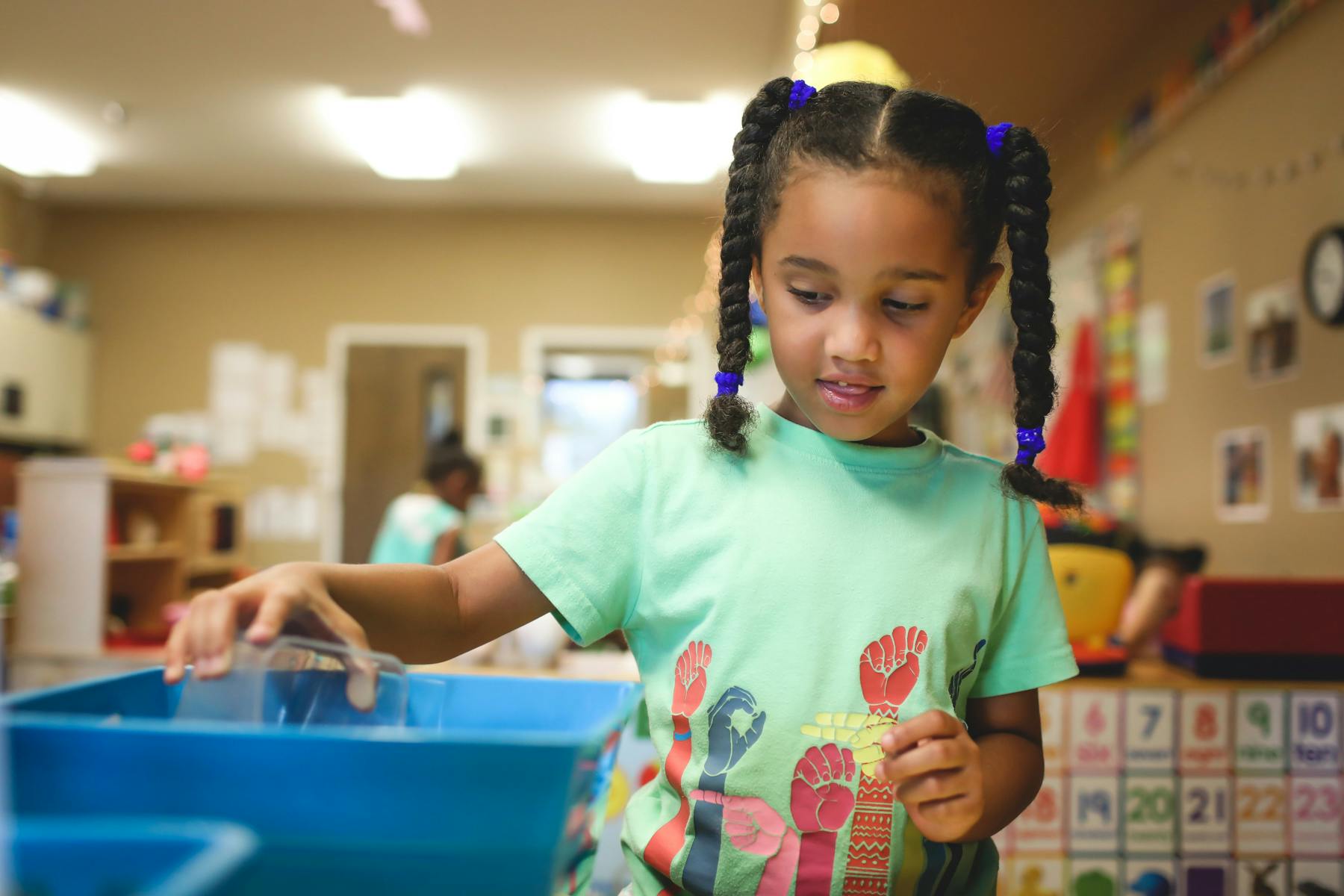 Child with braided pigtails reaching into a blue bin in a classroom
