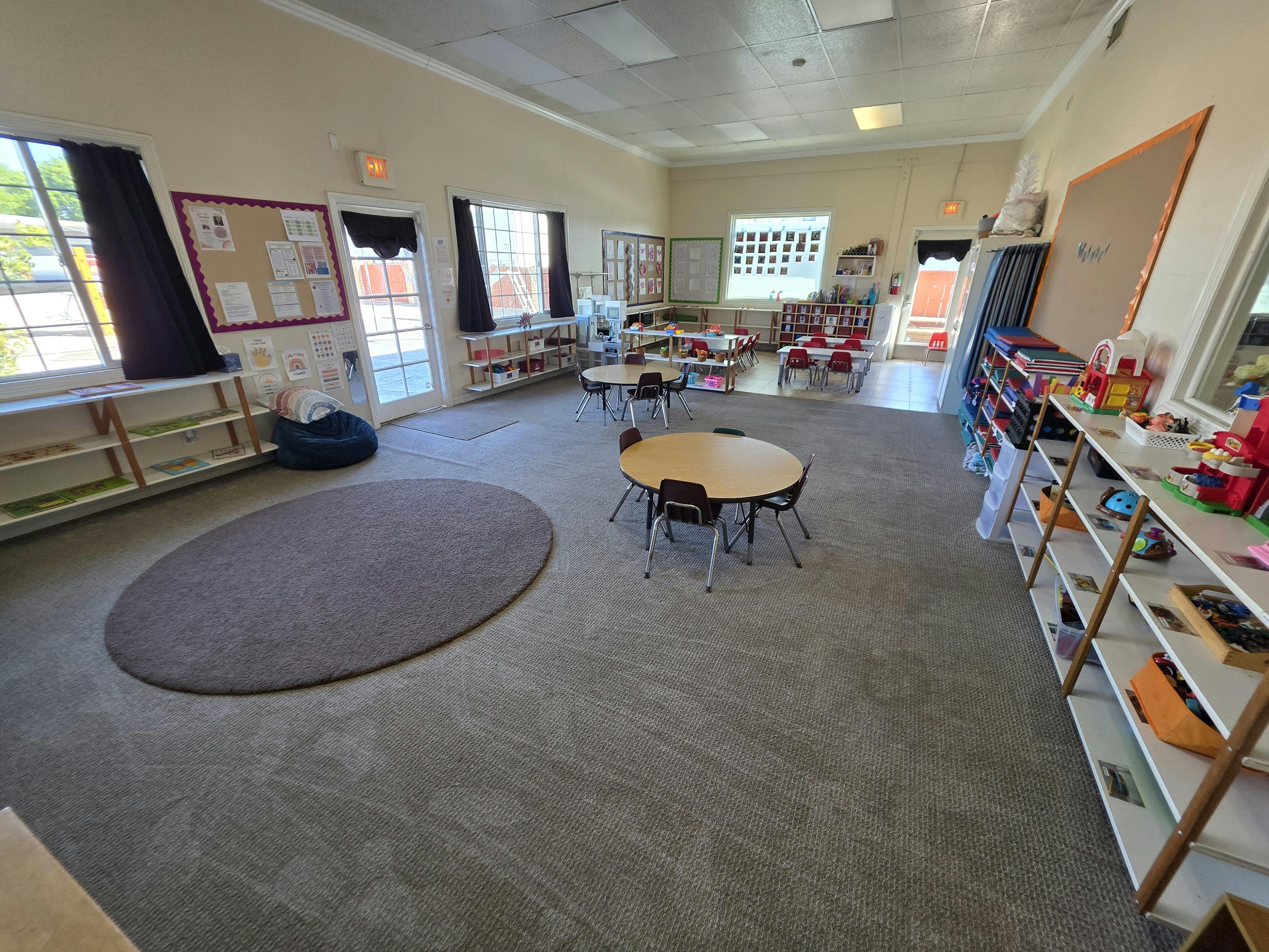 An empty classroom with small tables and chairs, shelves with toys, and bulletin boards