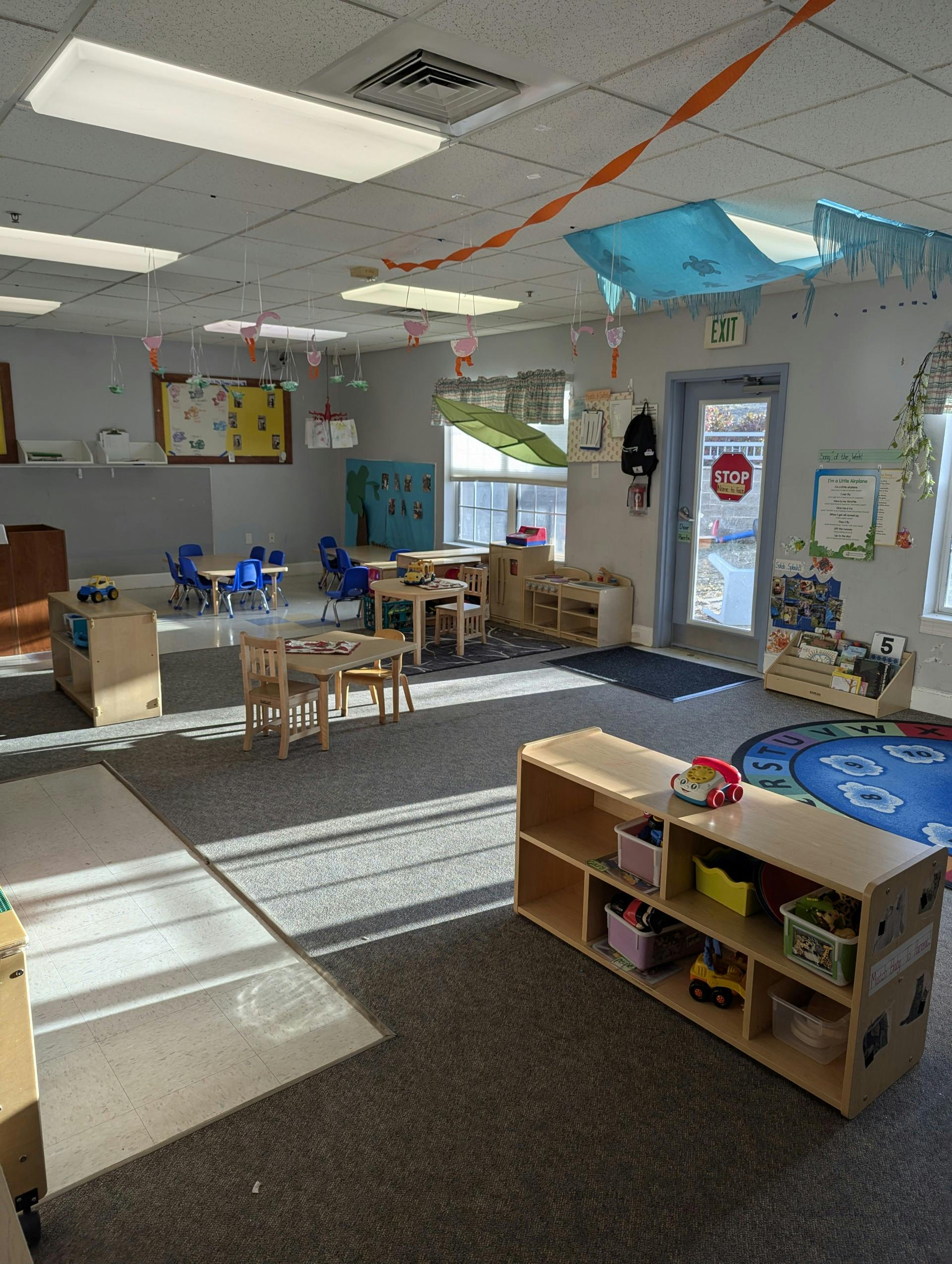 Classroom with tables, chairs, shelving units with toys, and decorations hanging from the ceiling
