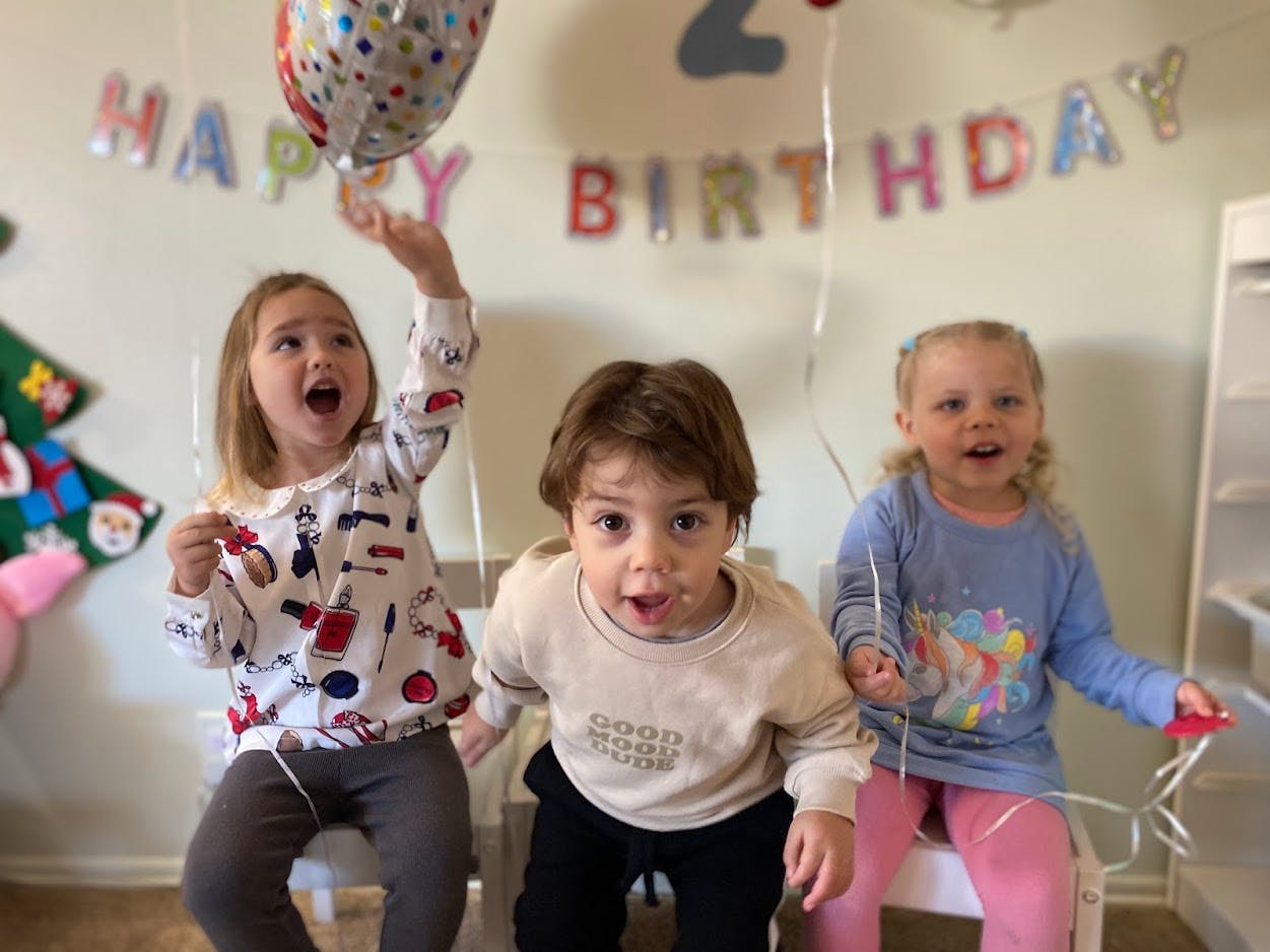 Three children sitting, with a \"Happy Birthday\" banner in the background