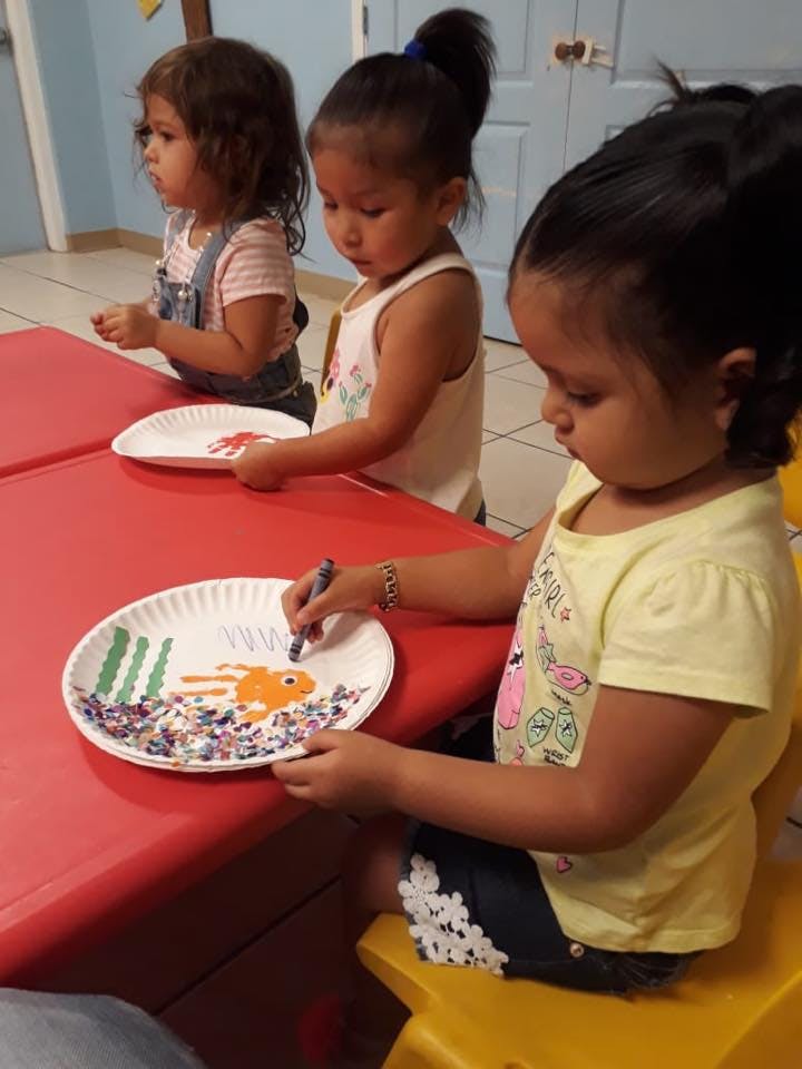 Three children at a table, one drawing on a paper plate with a marker