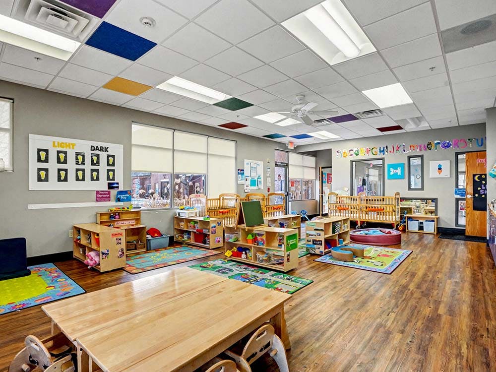 An empty daycare room with cribs, small tables, and colorful rugs on the floor