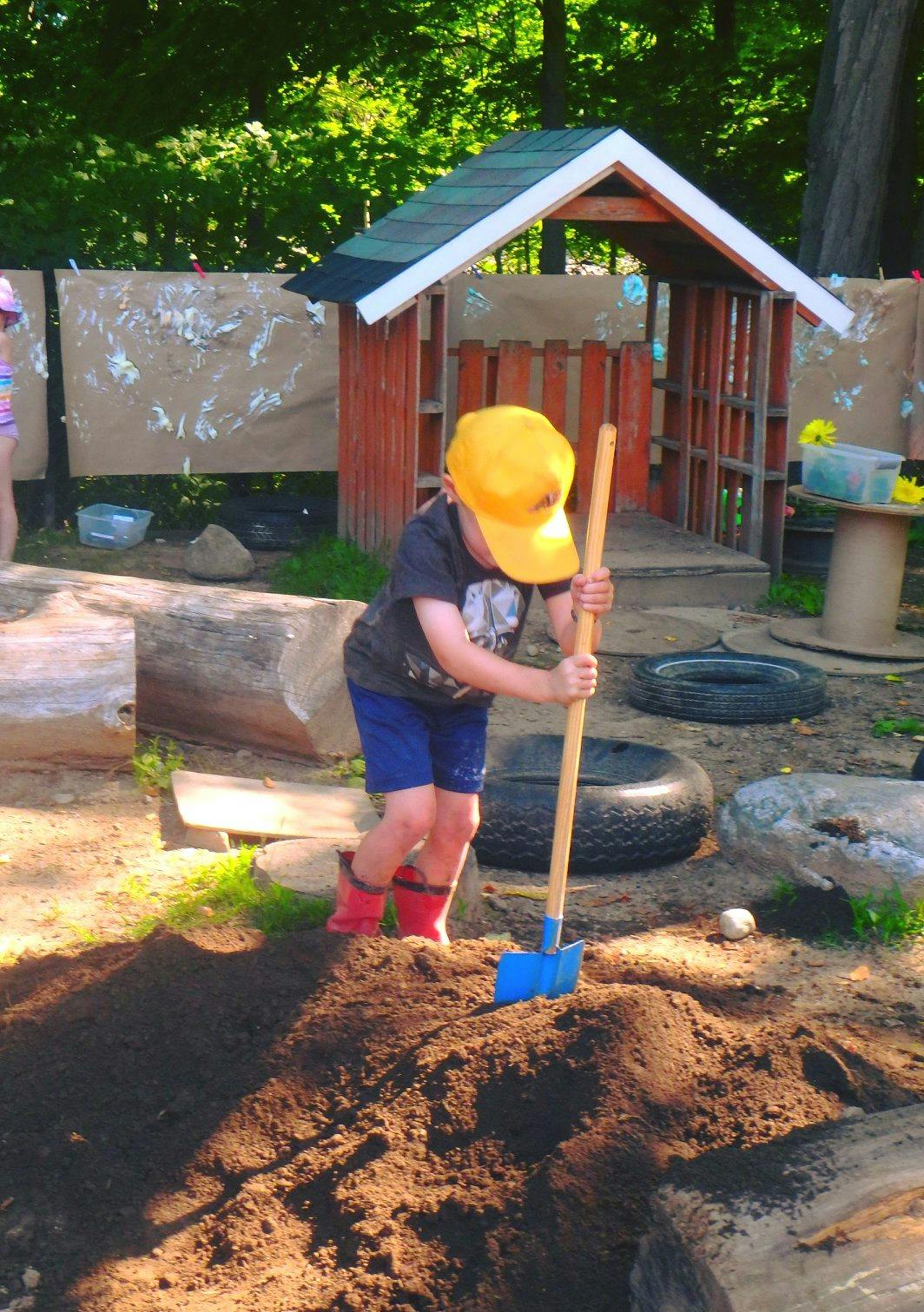 Child in a yellow hat and red boots digging in dirt with a shovel