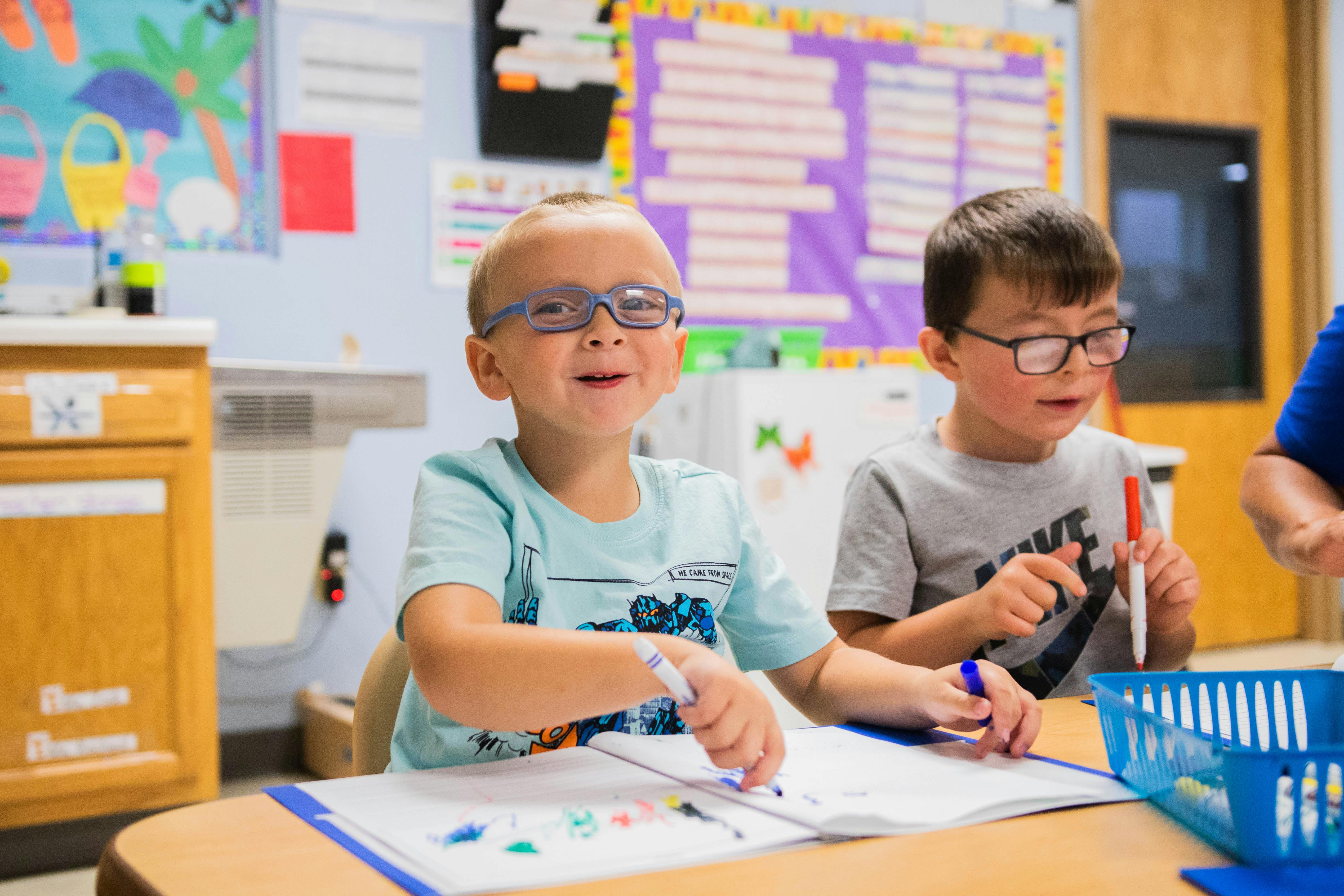 Two children with markers and paper at a table in a classroom
