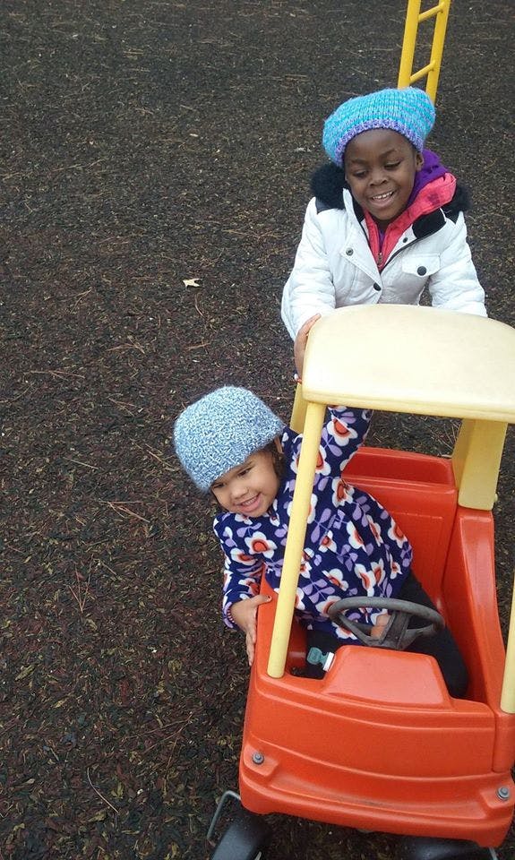 Child in a toy car with another child standing behind