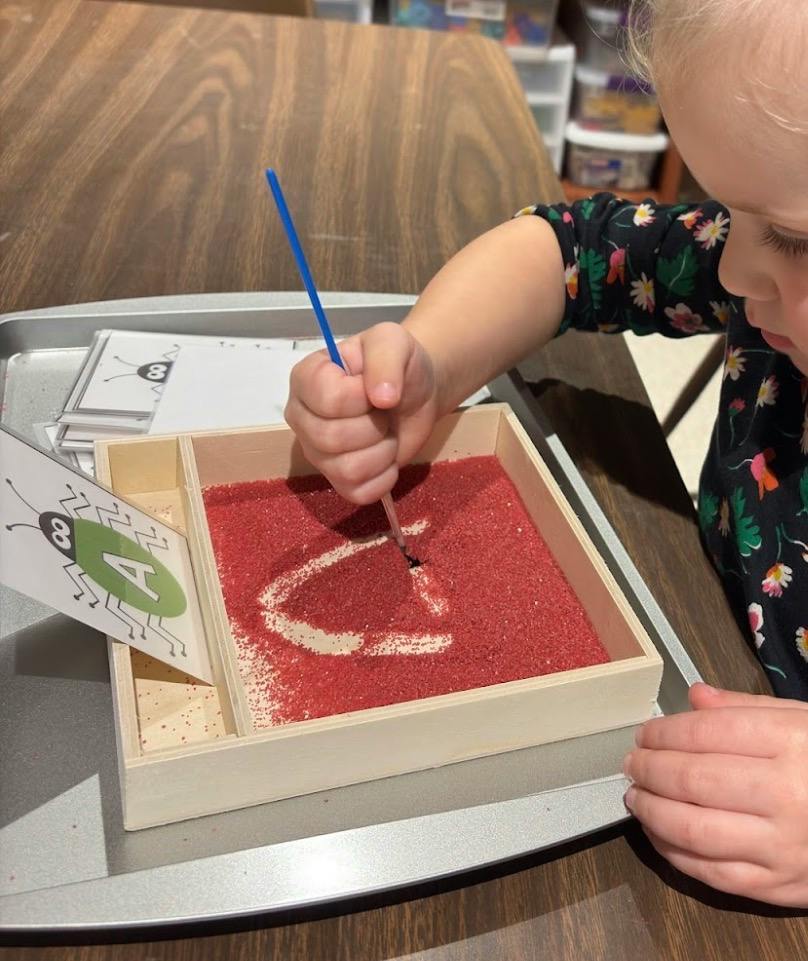 Child using a paintbrush to trace the letter A in a tray of red sand