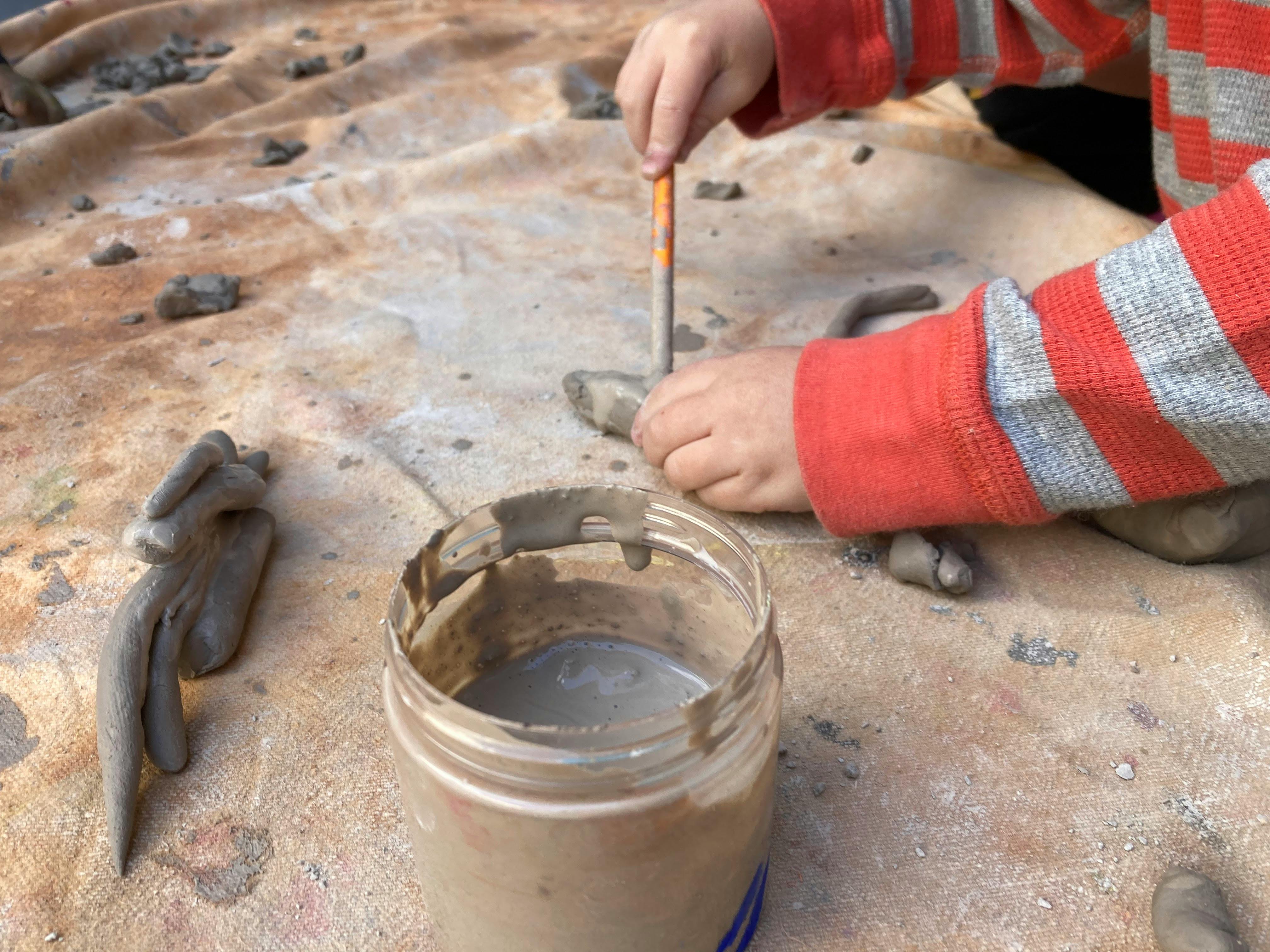 Child using a tool to shape clay next to a container of slip