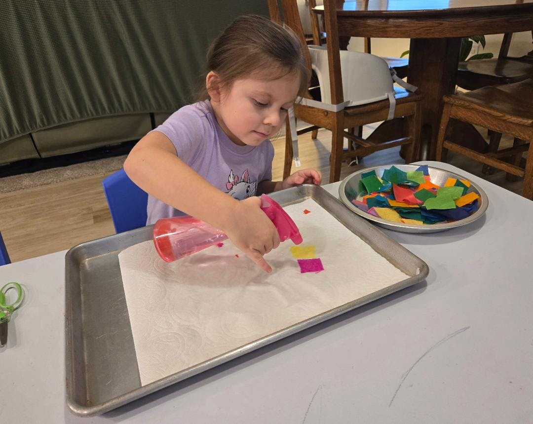 Child spraying paper with a bottle while small squares of colorful paper are on it