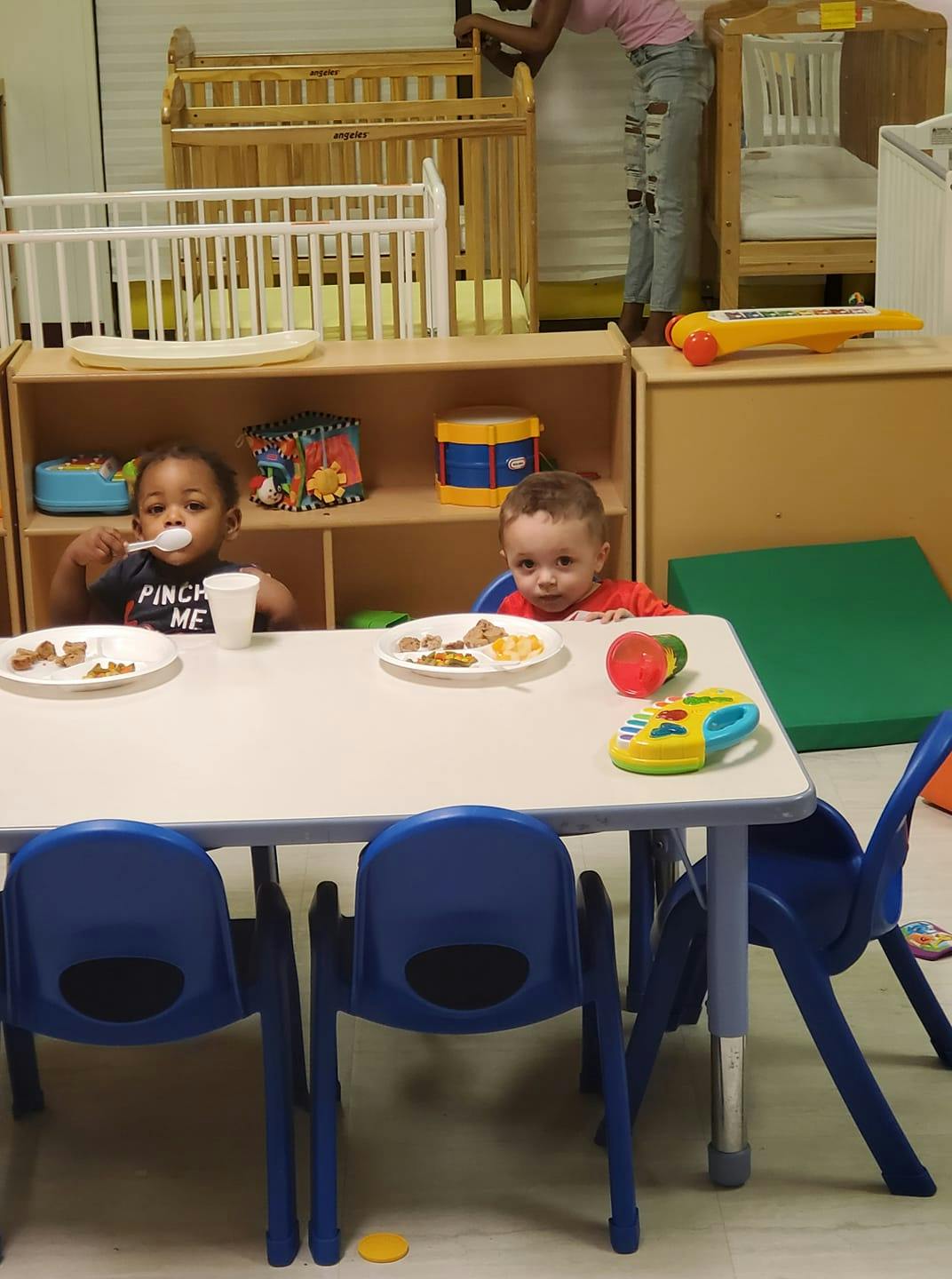 Two children eating at a table in a room with cribs
